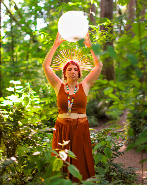 A woman with a glowing halo around her head, holding a glowing white orb above her head in a lush green forest.