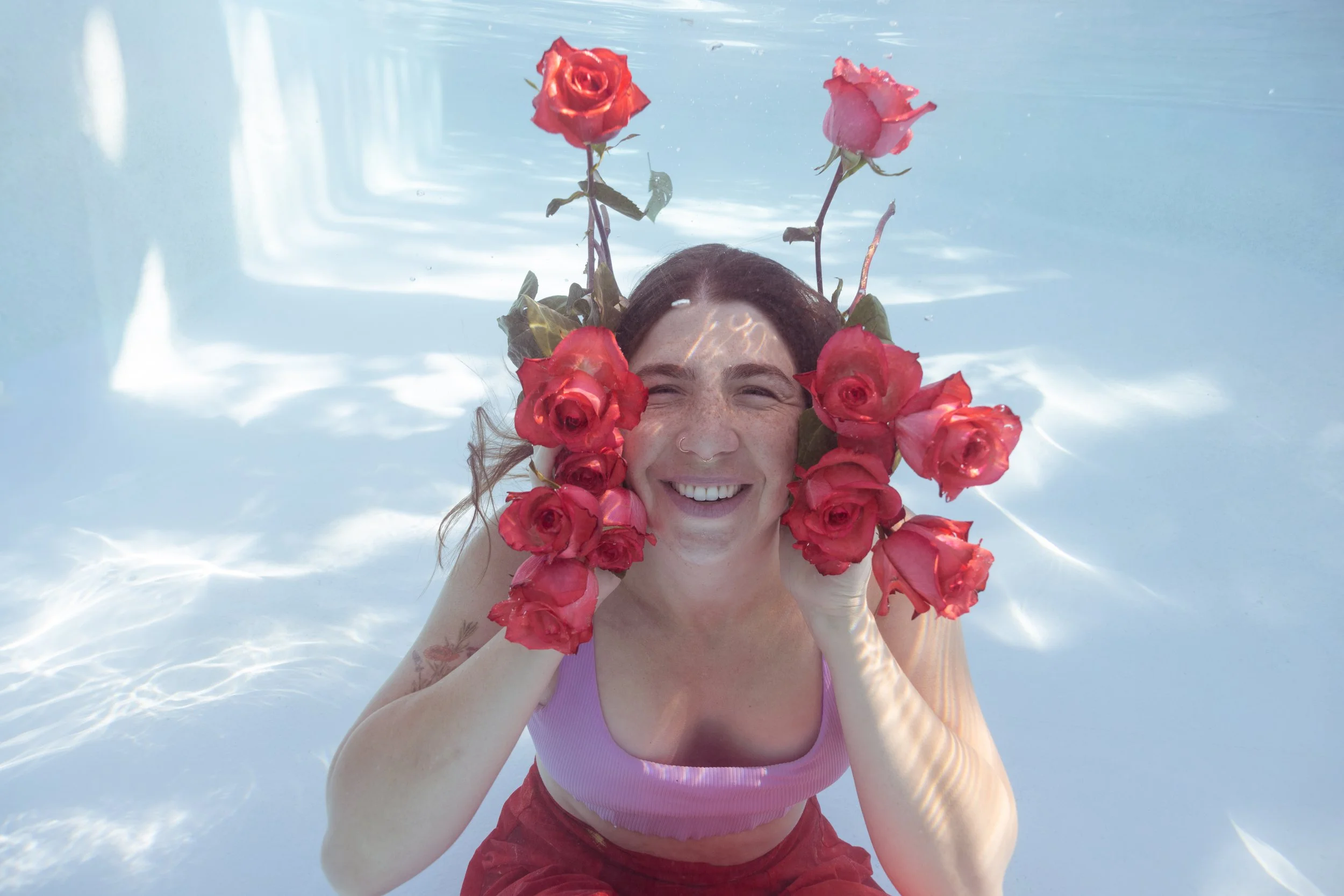 A woman smiling in a swimming pool with pink and red roses adorning her head and shoulders.