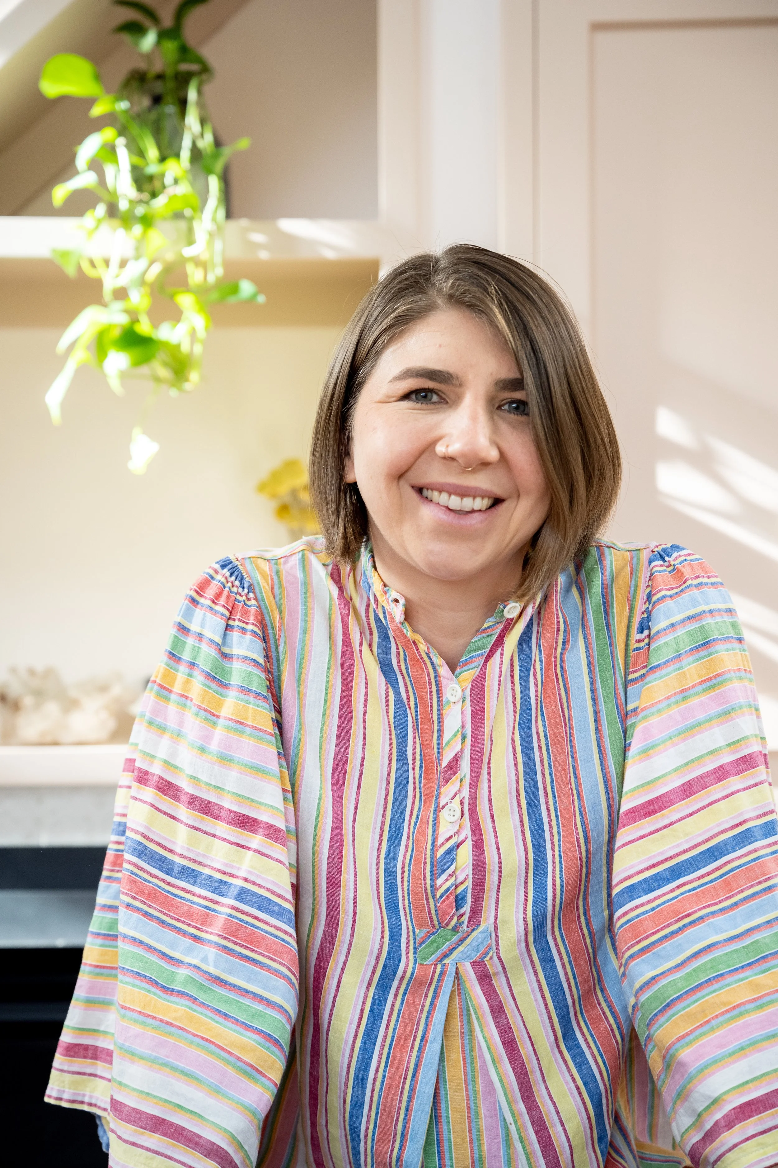 A young woman with shoulder-length brown hair and blue eyes sitting at a table in a room with natural lighting, wearing a colorful striped blouse.