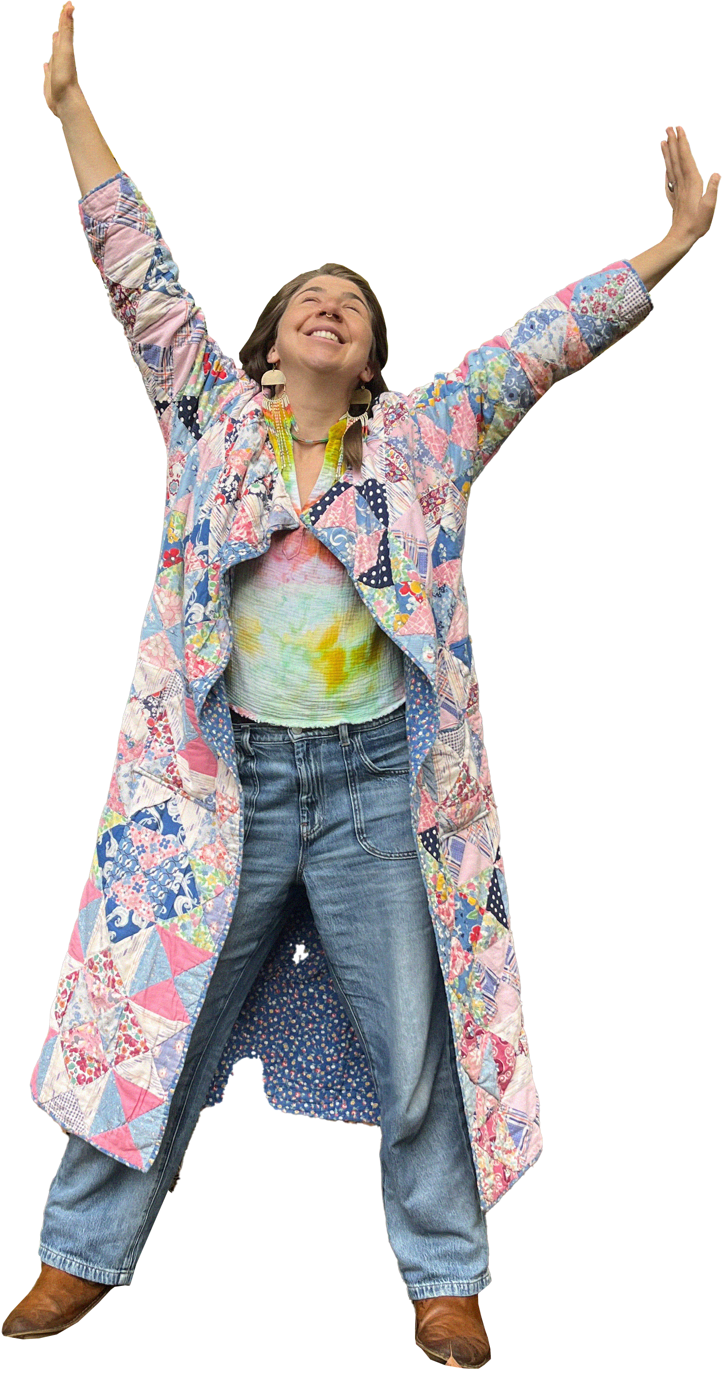 A woman in a colorful patchwork quilt coat, tie-dye shirt, jeans, and cowboy boots, smiling with her arms raised.