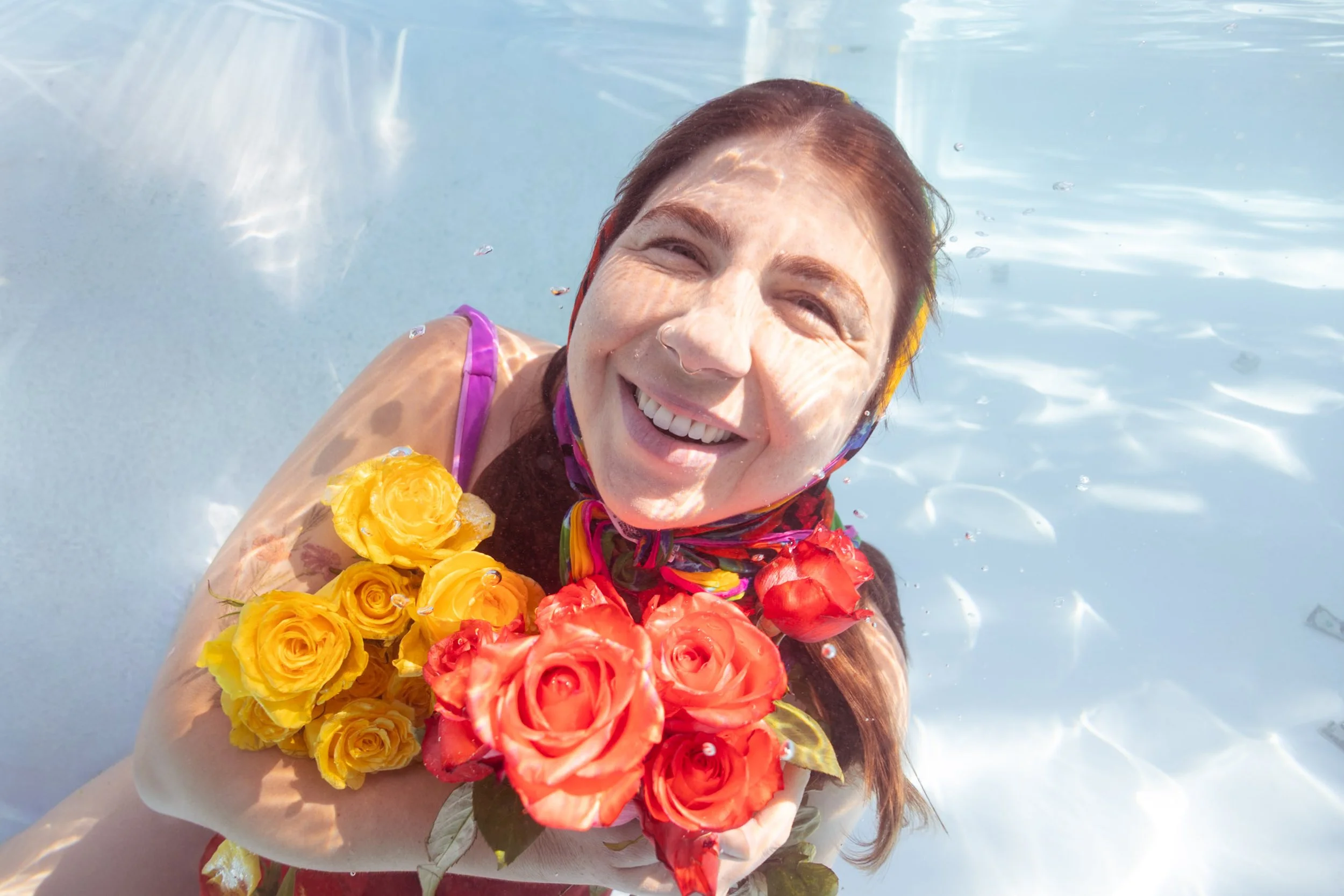 A smiling woman with red hair is floating in a pool, wearing a colorful swimsuit and holding a bouquet of yellow and orange roses.