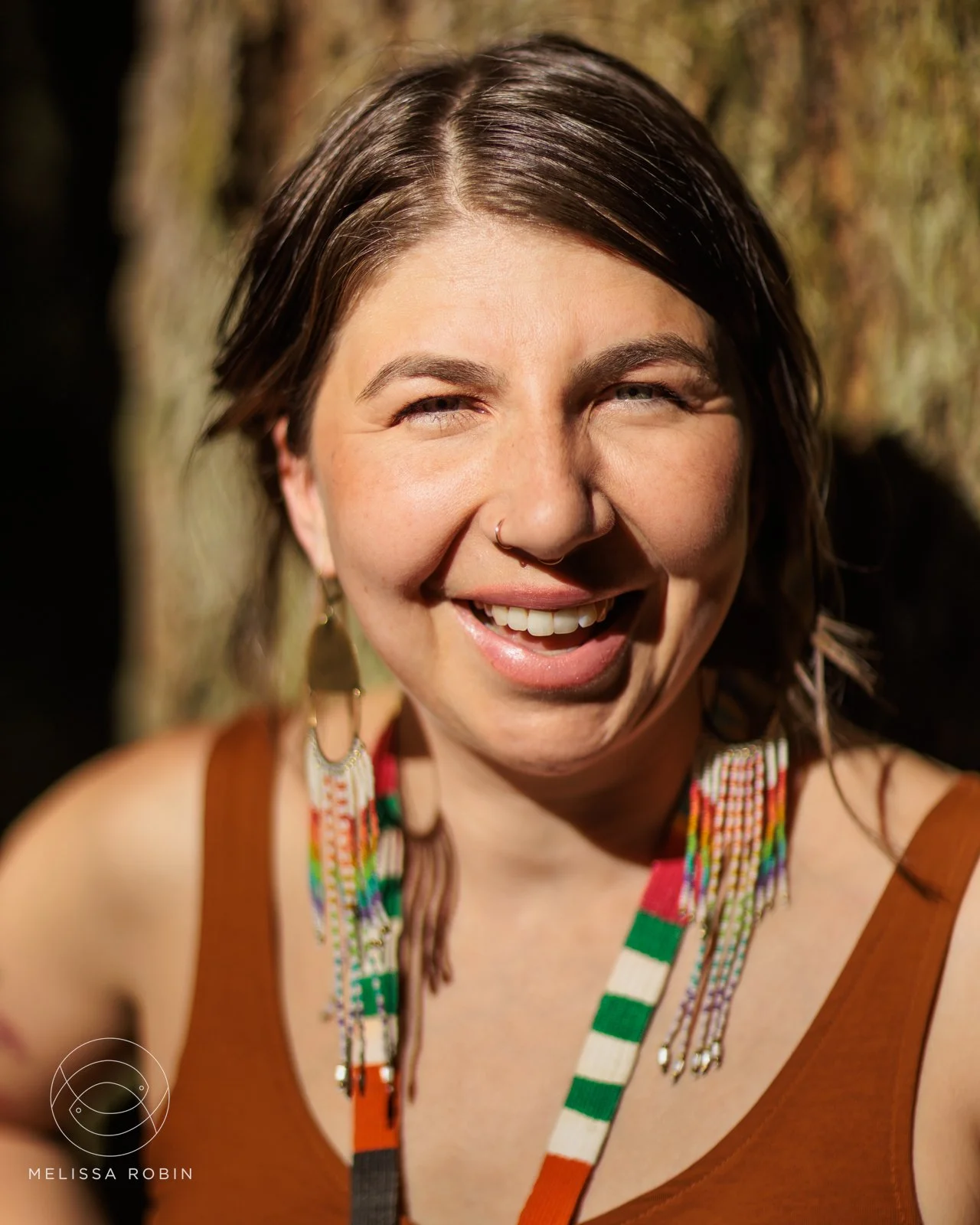 A young woman with short brown hair, smiling and squinting in sunlight, wearing a brown tank top and colorful beaded necklaces, standing outdoors with a tree trunk in the background.