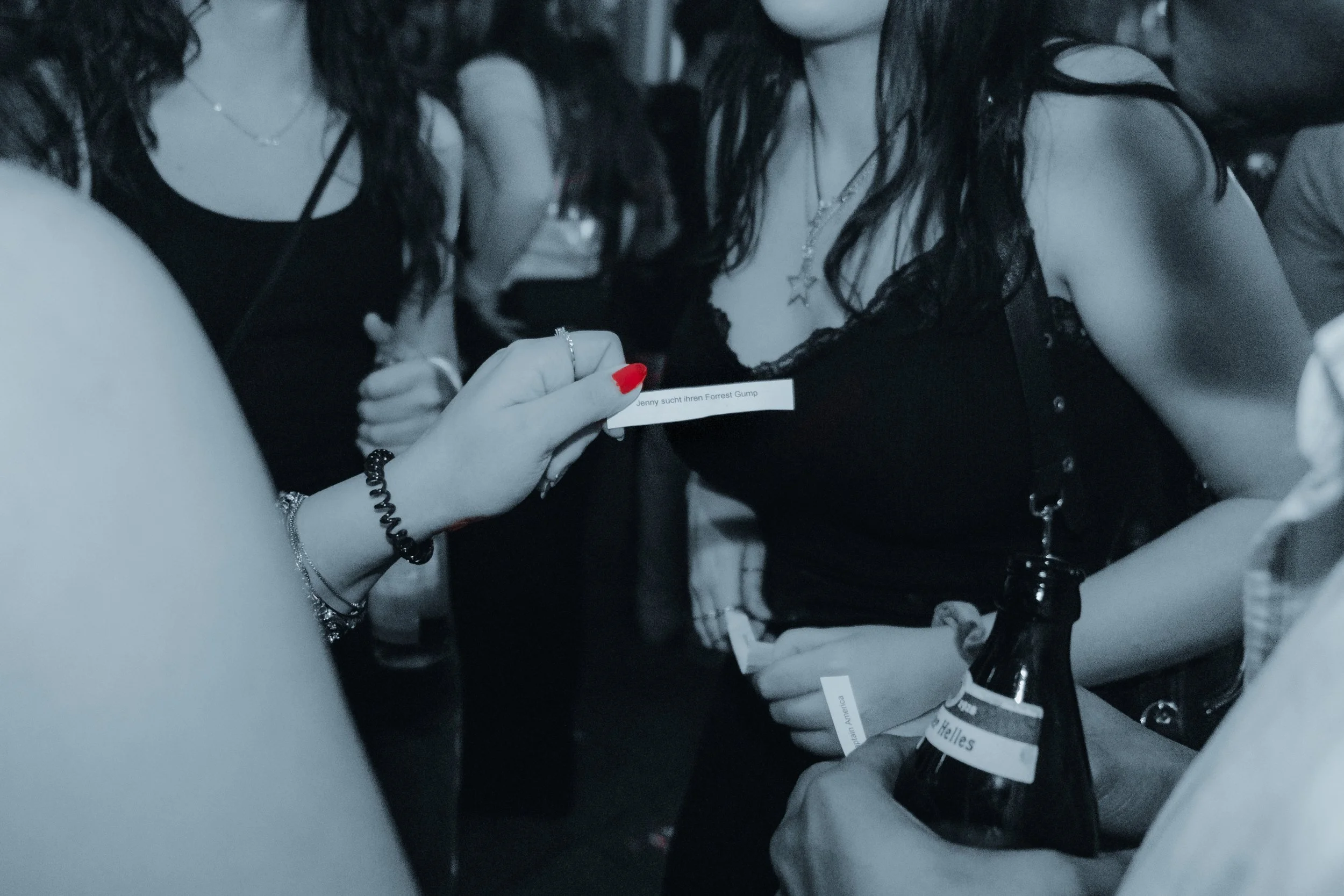 Four women standing closely together, dressed in black clothing, with one woman holding a small piece of paper. They are wearing necklaces and bracelets, and some have long hair. The scene appears to be in a dimly lit environment.