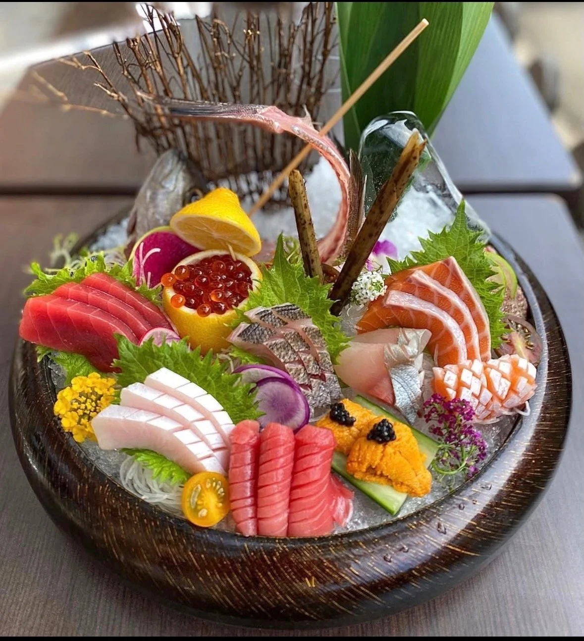 A large wooden bowl filled with various types of fresh sushi and sashimi arranged on ice, garnished with herbs, vegetables, lemon, and colorful edible flowers.