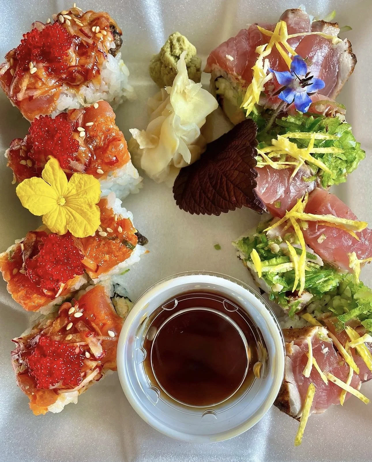 Assorted sushi rolls with various toppings, including fish, vegetables, flowers, and a small bowl of soy sauce or dipping sauce. Sushi by Devin.