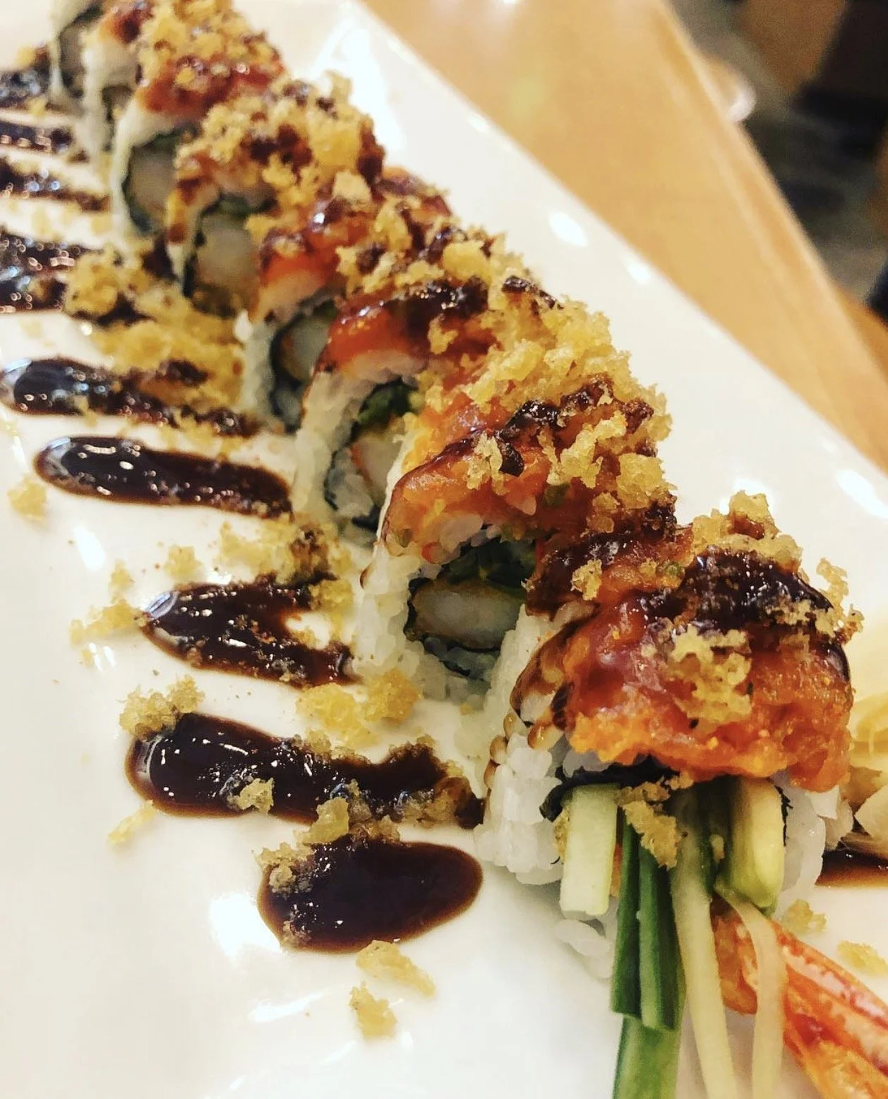 Sushi roll topped with tempura flakes, drizzled with eel sauce, served on a white plate with additional eel sauce. Sushi by Devin.