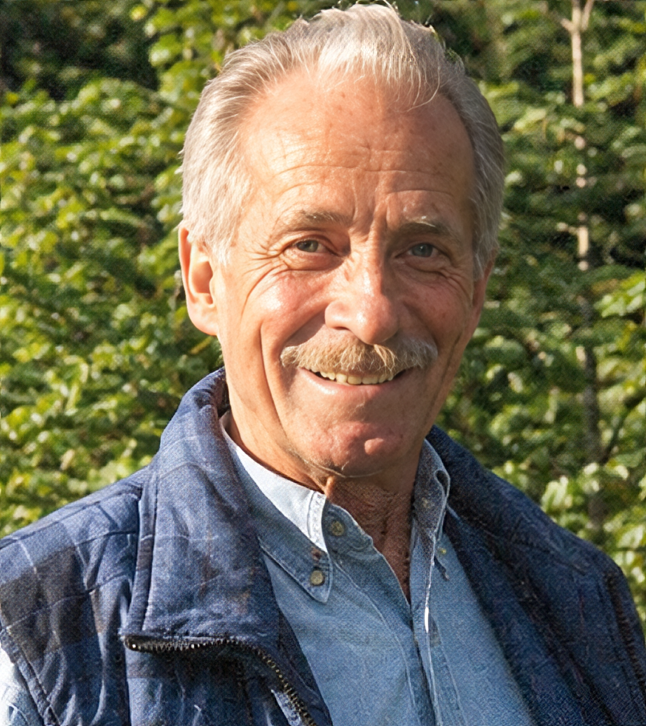 Un homme âgé souriant portant une chemise en jean et une veste en nylon, avec un fond de verdure dense.