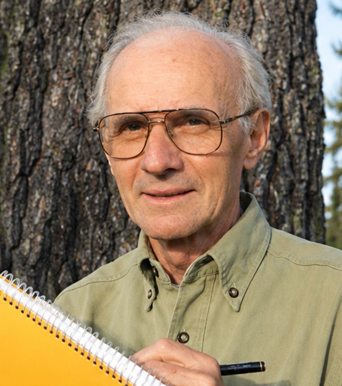 Un homme âgé portait des lunettes, une chemise bouton-ouverte et tient un carnet jaune avec un stylo, devant un arbre en forêt.