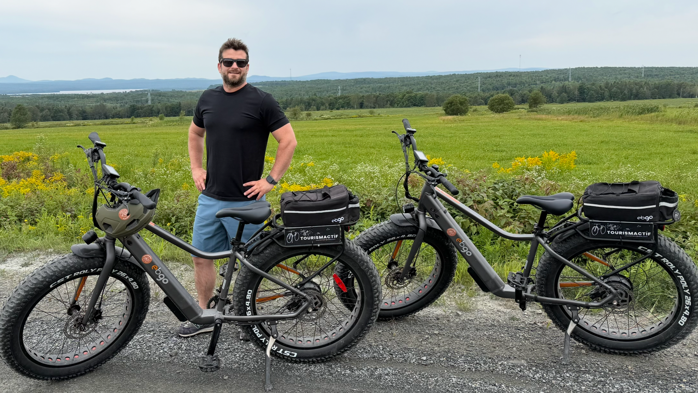 Un homme souriant en sunglasses, portant un t-shirt noir et un short bleu, se tient entre deux vélos électriques équipés de sacs de voyage, dans un paysage rural avec champs verts et montagnes en arrière-plan.