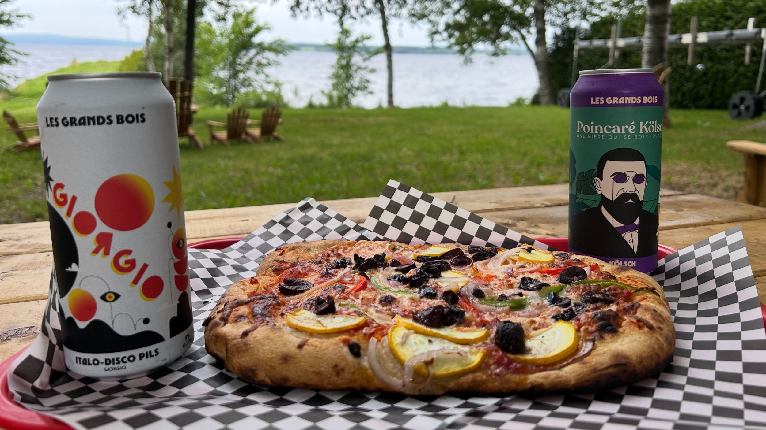 Pizza avec des légumes et de la viande sur une table en bois, deux canettes de bière, une dans chaque main, avec un décor naturel de arbres près d'un lac.