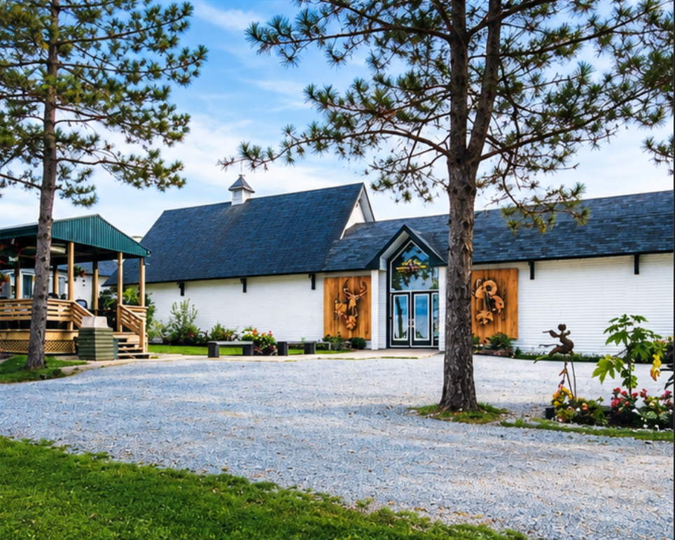 Une ferme ou une grange blanche en bois avec des décorations en bois au mur, entourée d'arbres, avec une allée en gravier, un porche en bois, et des sculptures de jardin.