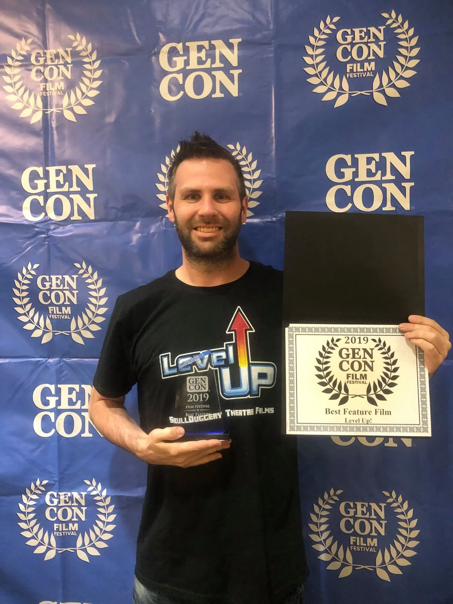 A man smiling and holding two awards in front of a blue backdrop with 'Gen Con Film Festival' logos. One award reads 'Best Feature Film' and the other 'Level Up!'