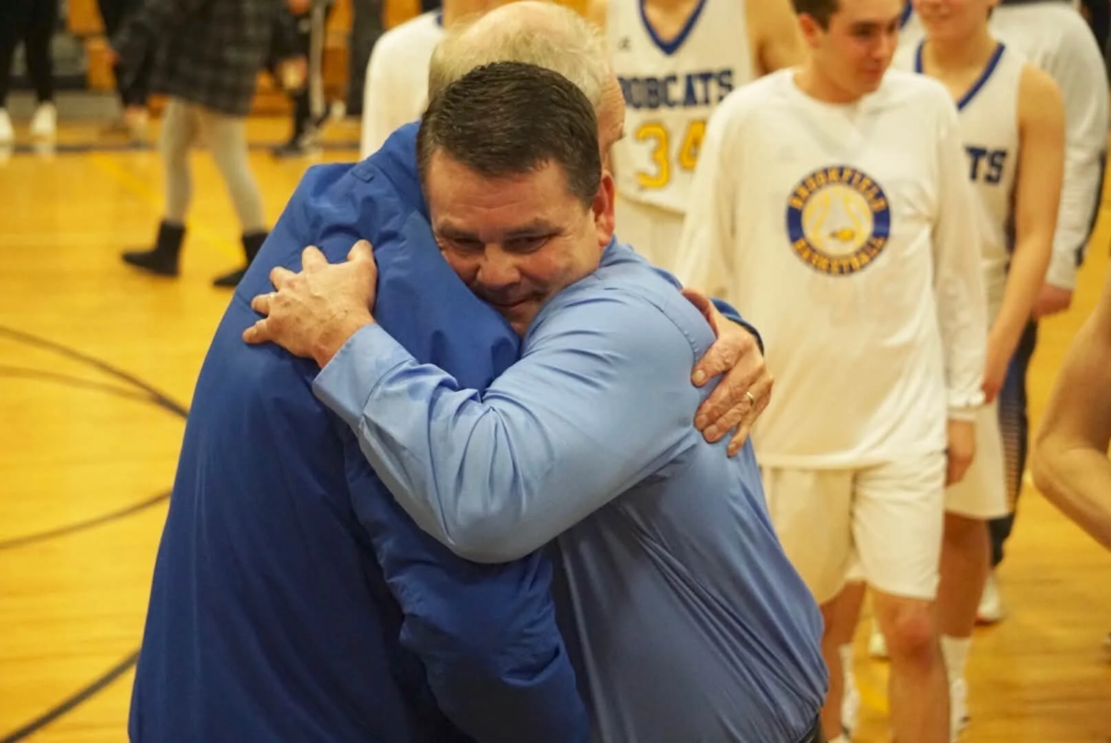 Two adults hugging in celebration on a basketball court with a team wearing white jerseys in the background.