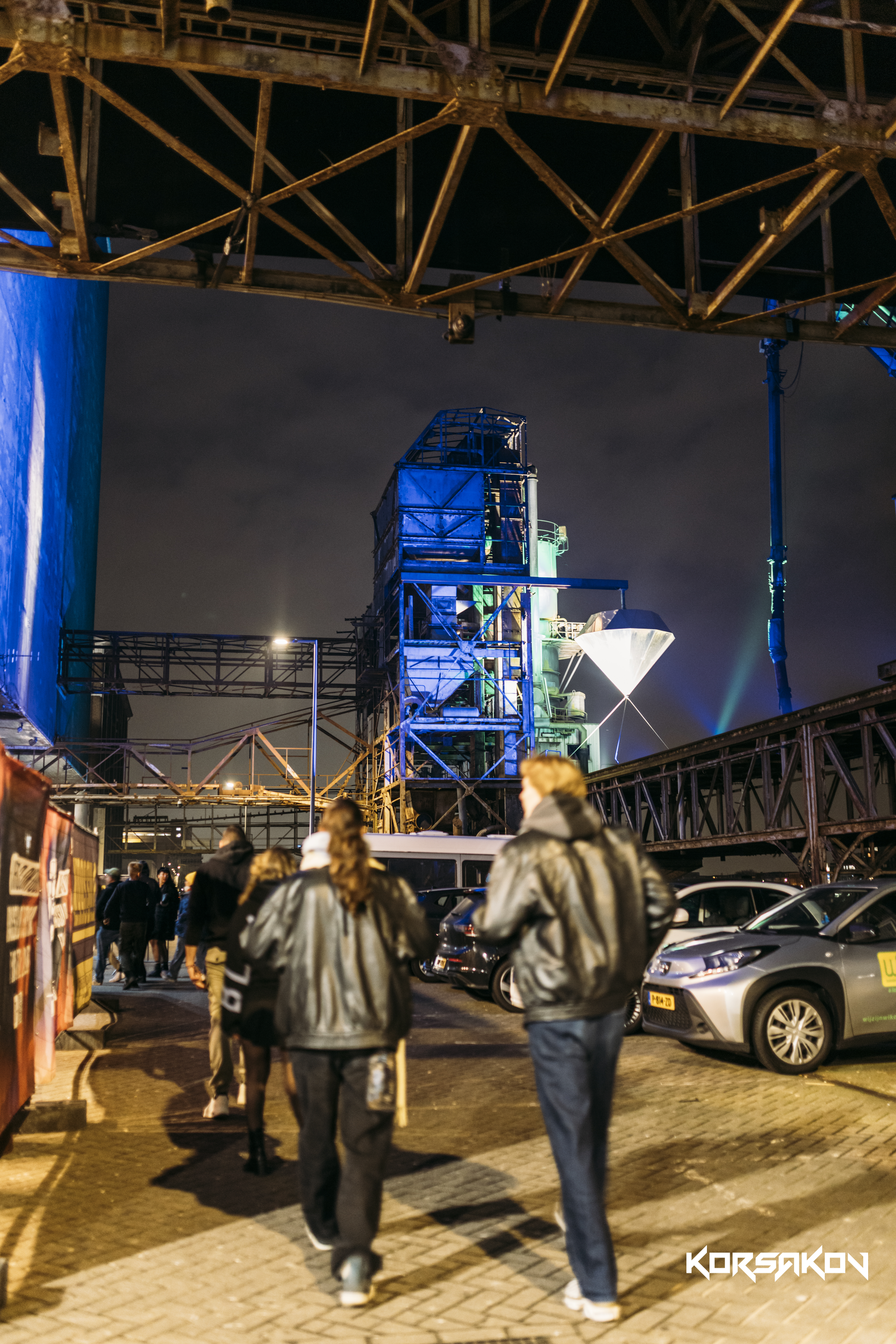 Night scene of people walking near parked cars with industrial structures and metal framework overhead, illuminated by colored lights in the background.
