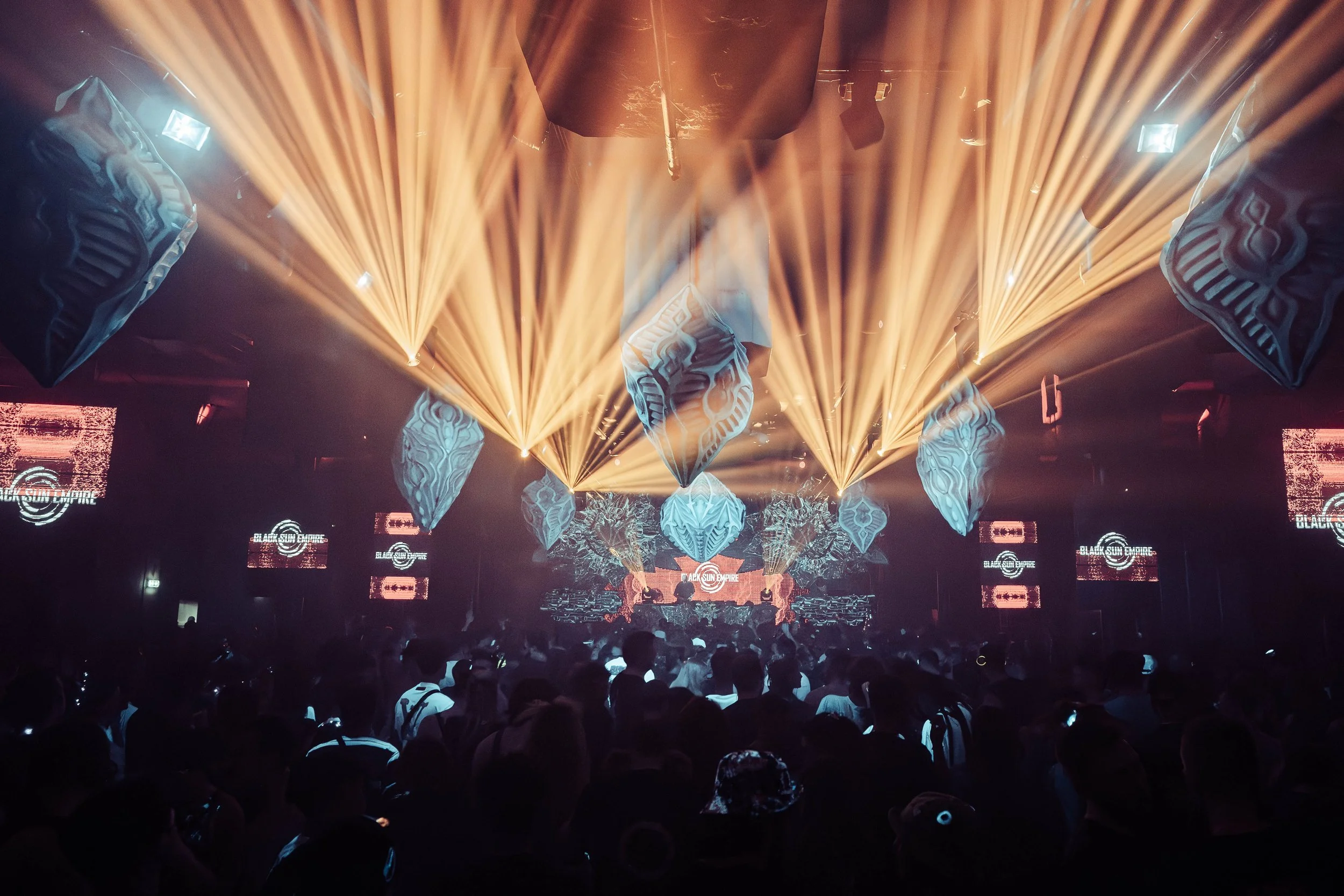A concert scene with yellow stage lights shining down, abstract decorative elements hanging from the ceiling, and a dark audience area with silhouettes of people watching the performance.