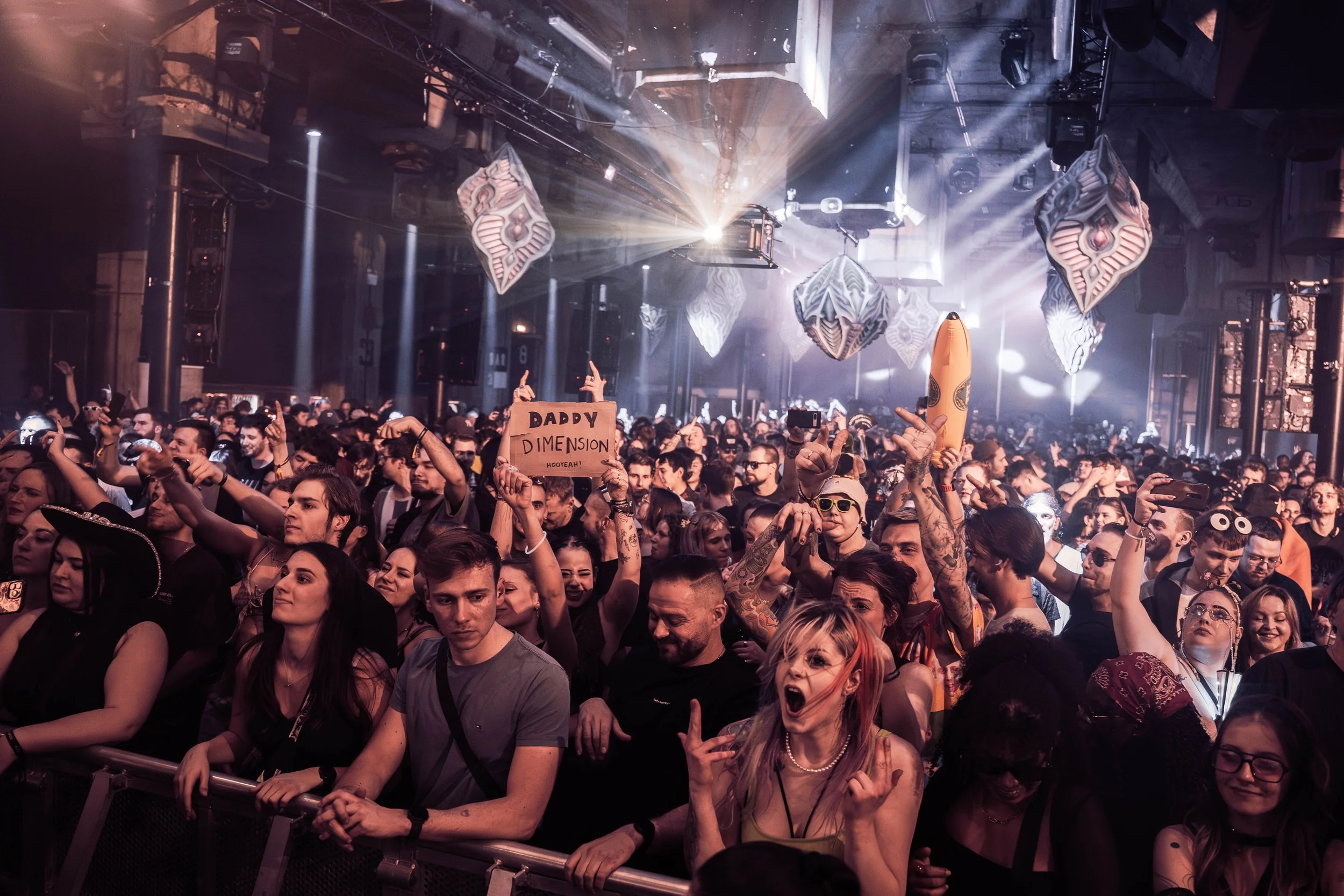 A large audience at a concert or dance event with people dancing, raising their hands, and holding signs. Bright stage lights and hanging geometric decorations are visible with a lively atmosphere.