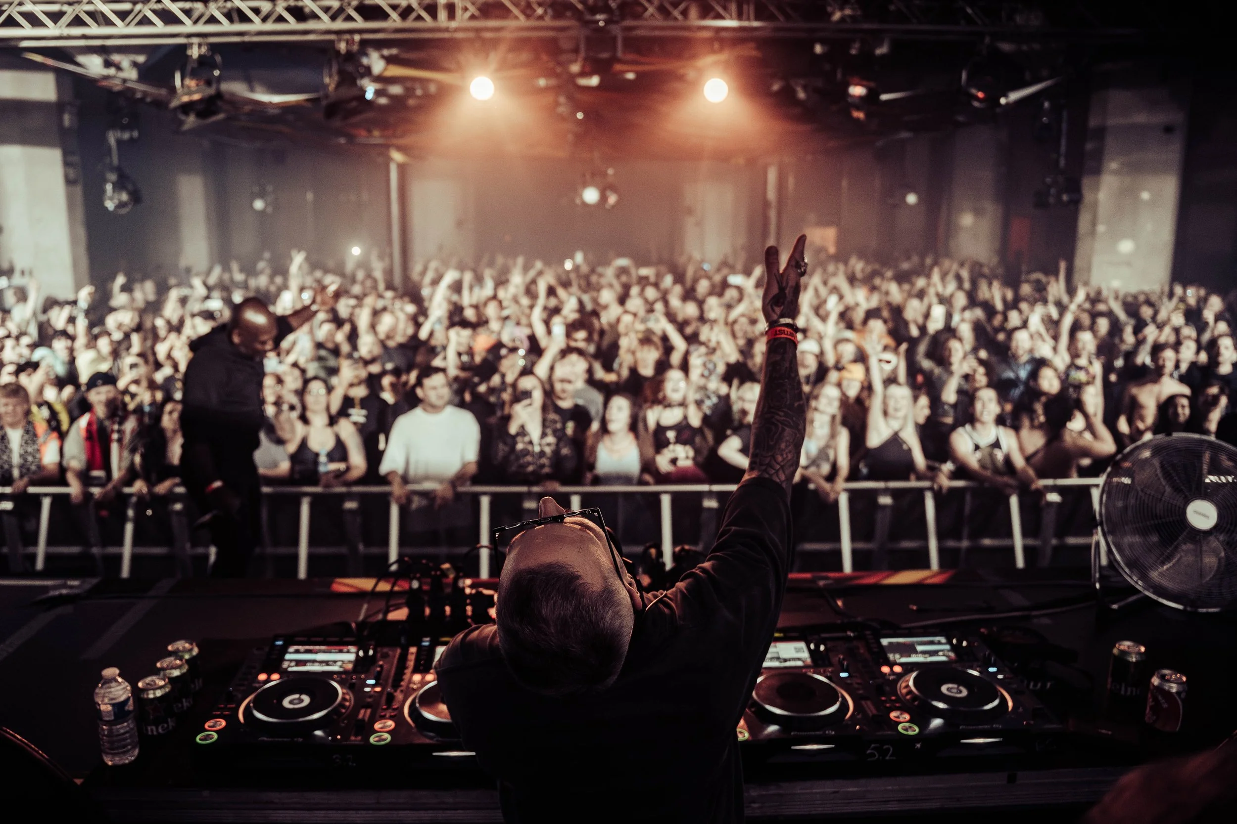 DJ performing at a concert with a crowd in front of him. The photo is taken from behind the DJ's equipment, showing the crowd raising their hands and enjoying the music. The venue is dimly lit with stage lights overhead.