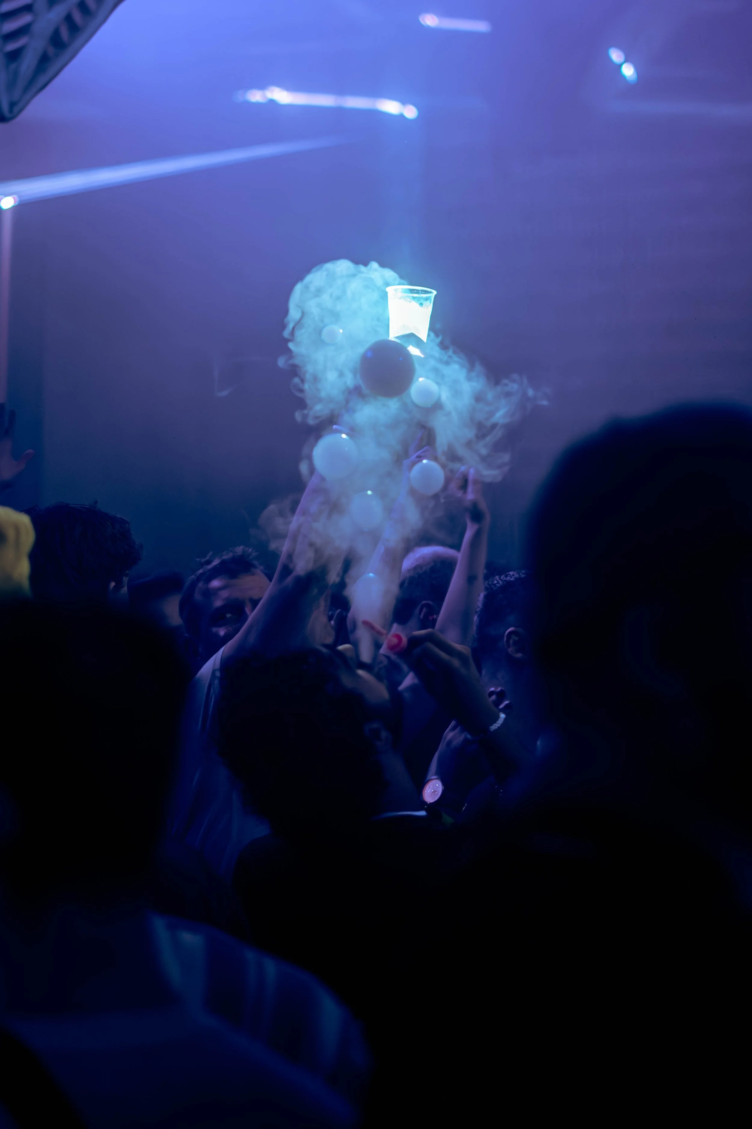 A crowd at a concert or nightclub with people raising their hands and holding drinks, illuminated by blue and purple stage lighting.