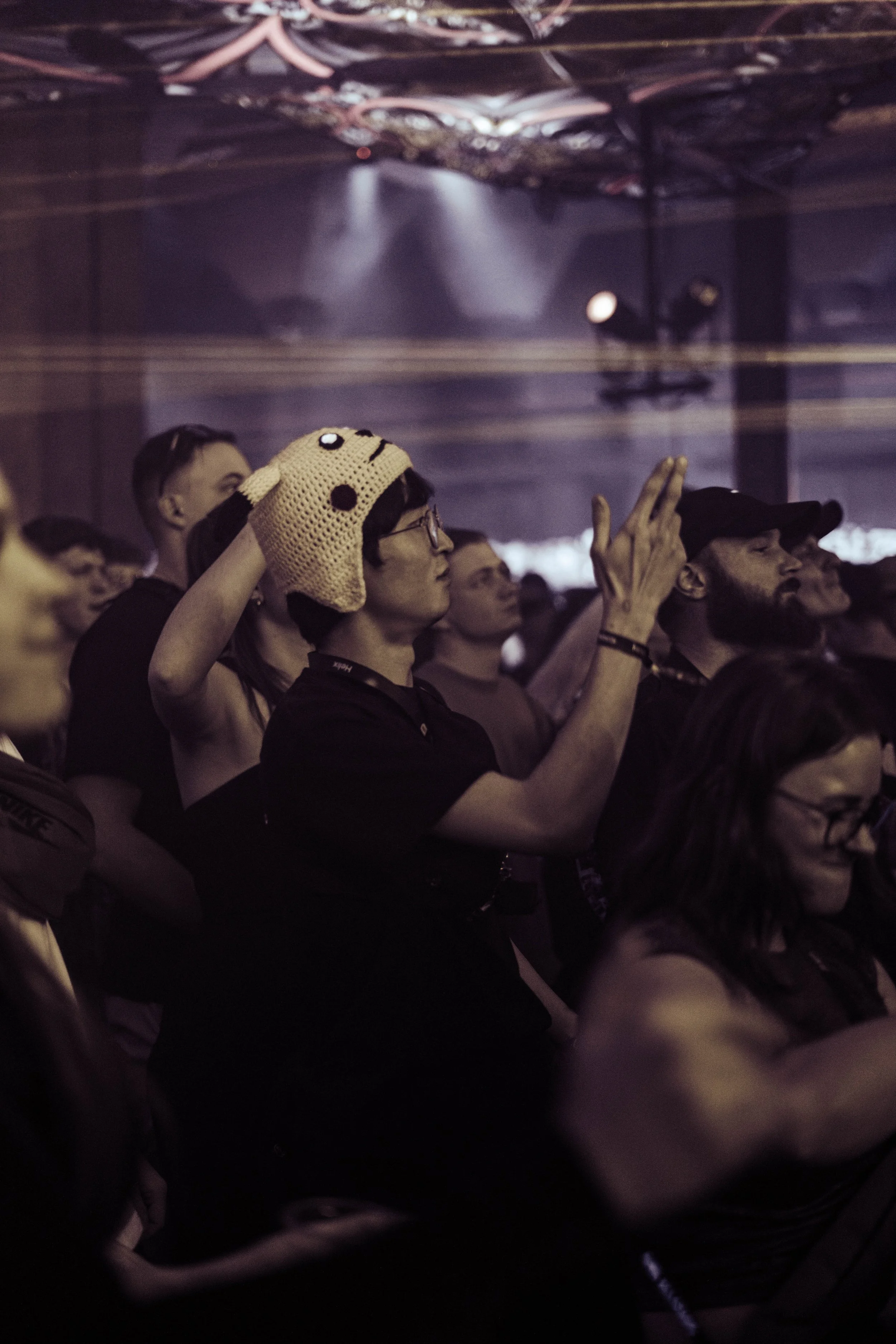 Audience at concert or event, some with hats and glasses, standing in dim lighting, facing stage or performance area.