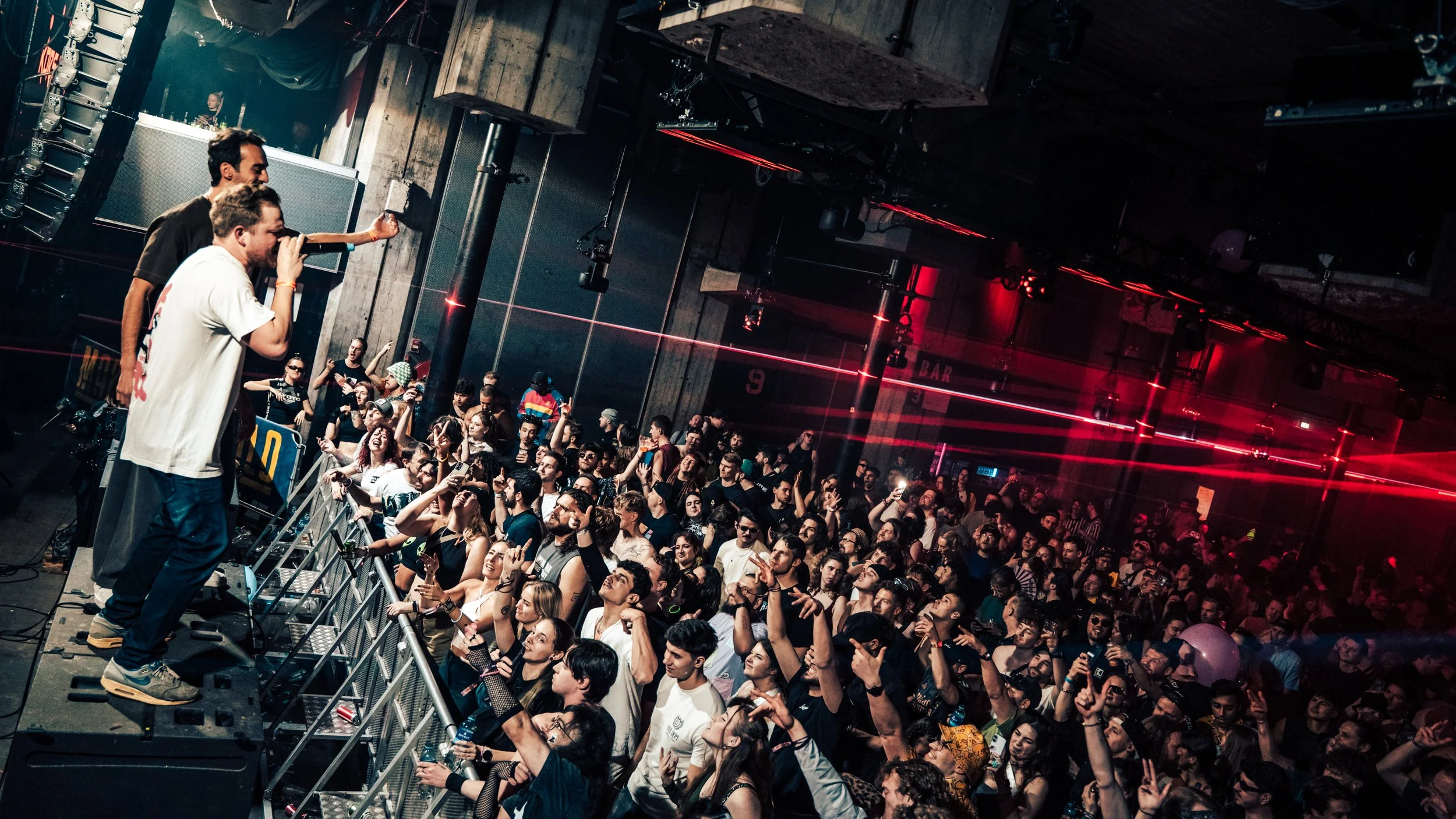 A DJ and an MC are performing at a music concert with a large crowd dancing and enjoying the show in a dark club with red and black lighting.