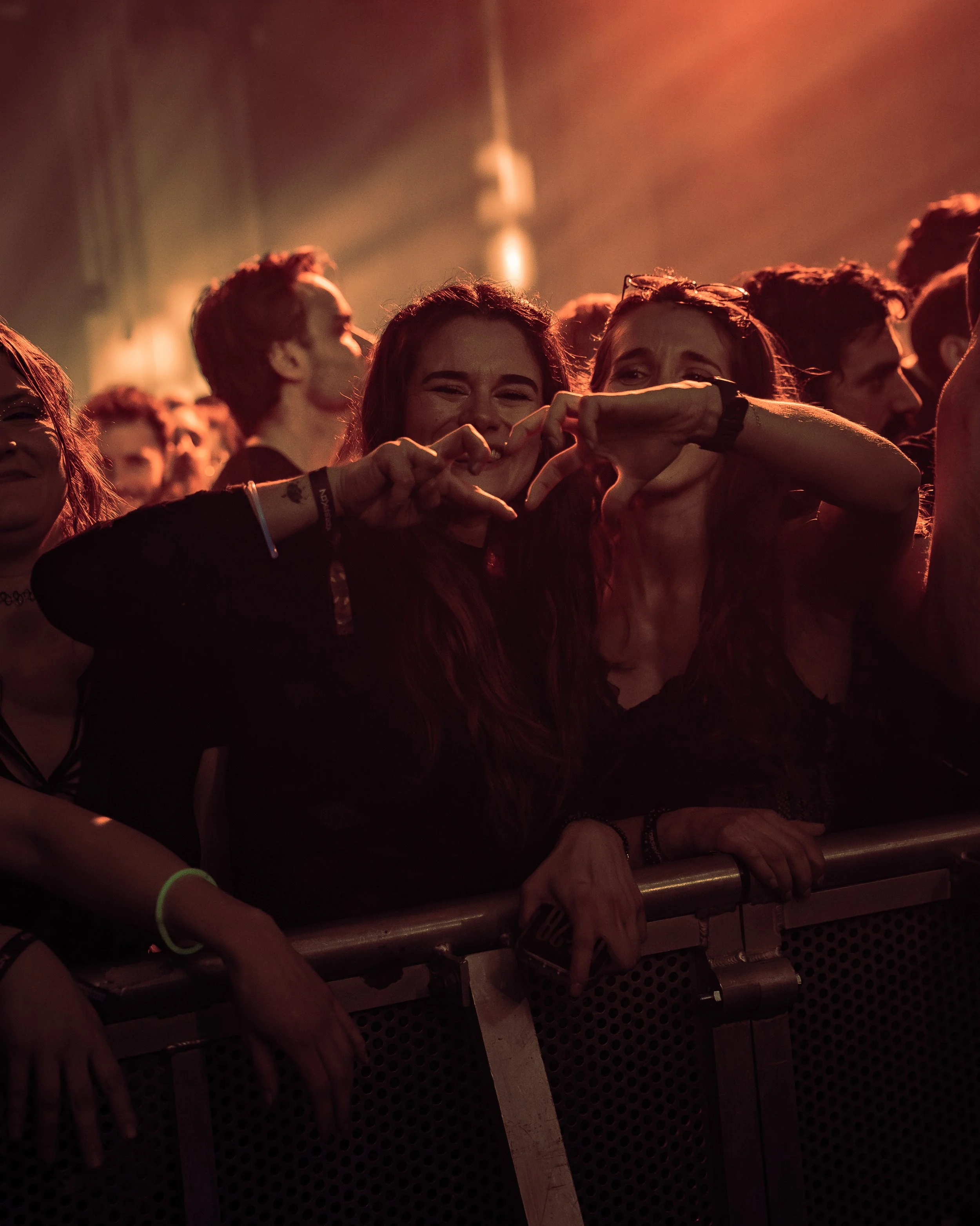 Crowd of people at concert or event, some smiling and making peace signs, with low lighting and warm tones.