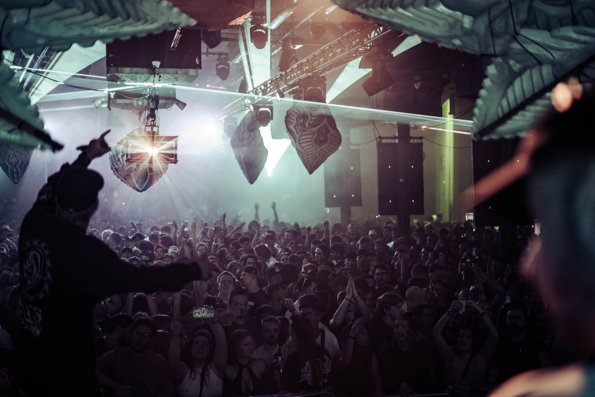 A large crowd at a concert or music event, with people raising their hands and facing the stage. The stage is visible in the background with lighting and sound equipment, and a performer is seen on stage.