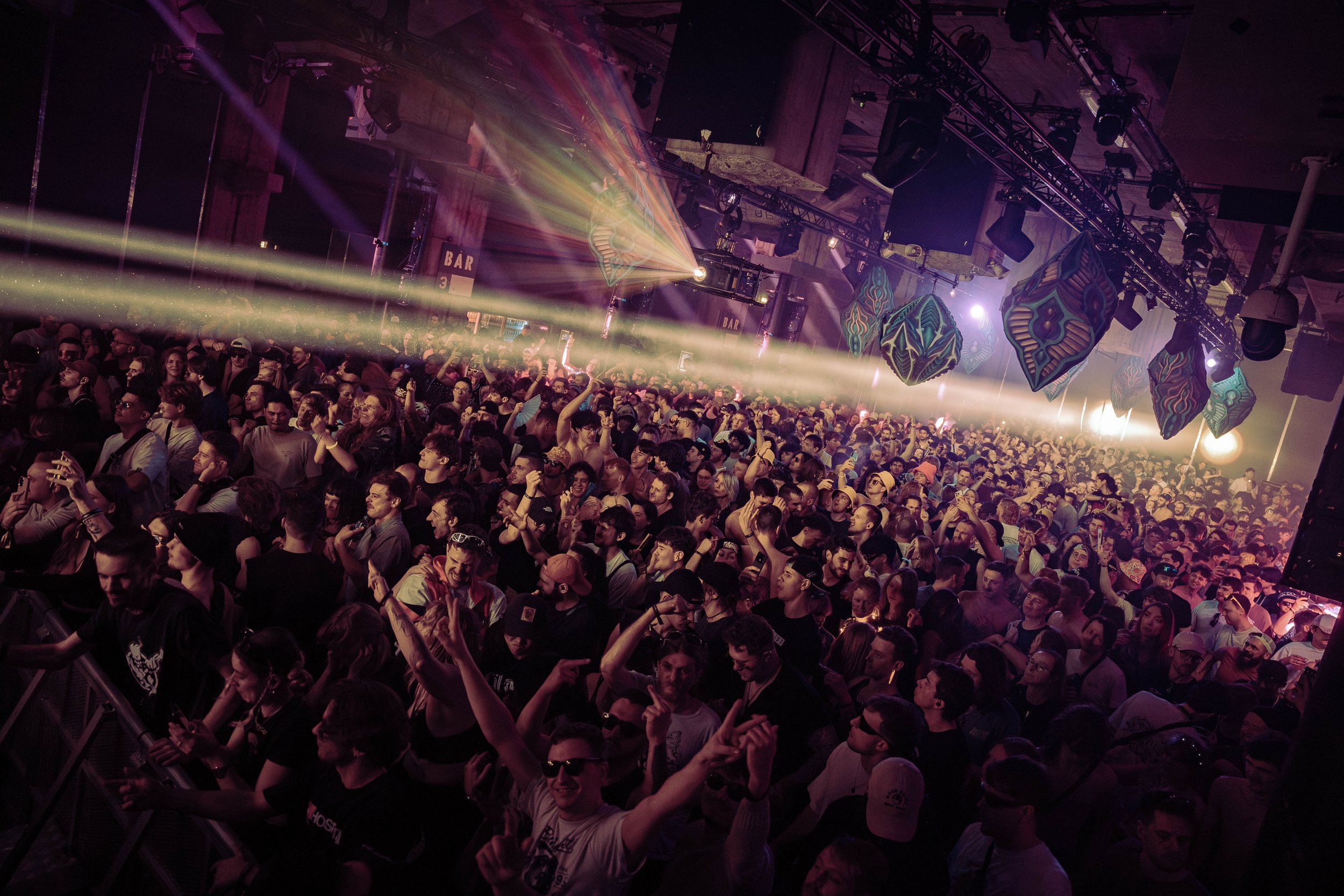 A crowded indoor music concert venue with people dancing and enjoying the performance, colorful stage lights, and decorations hanging from the ceiling.