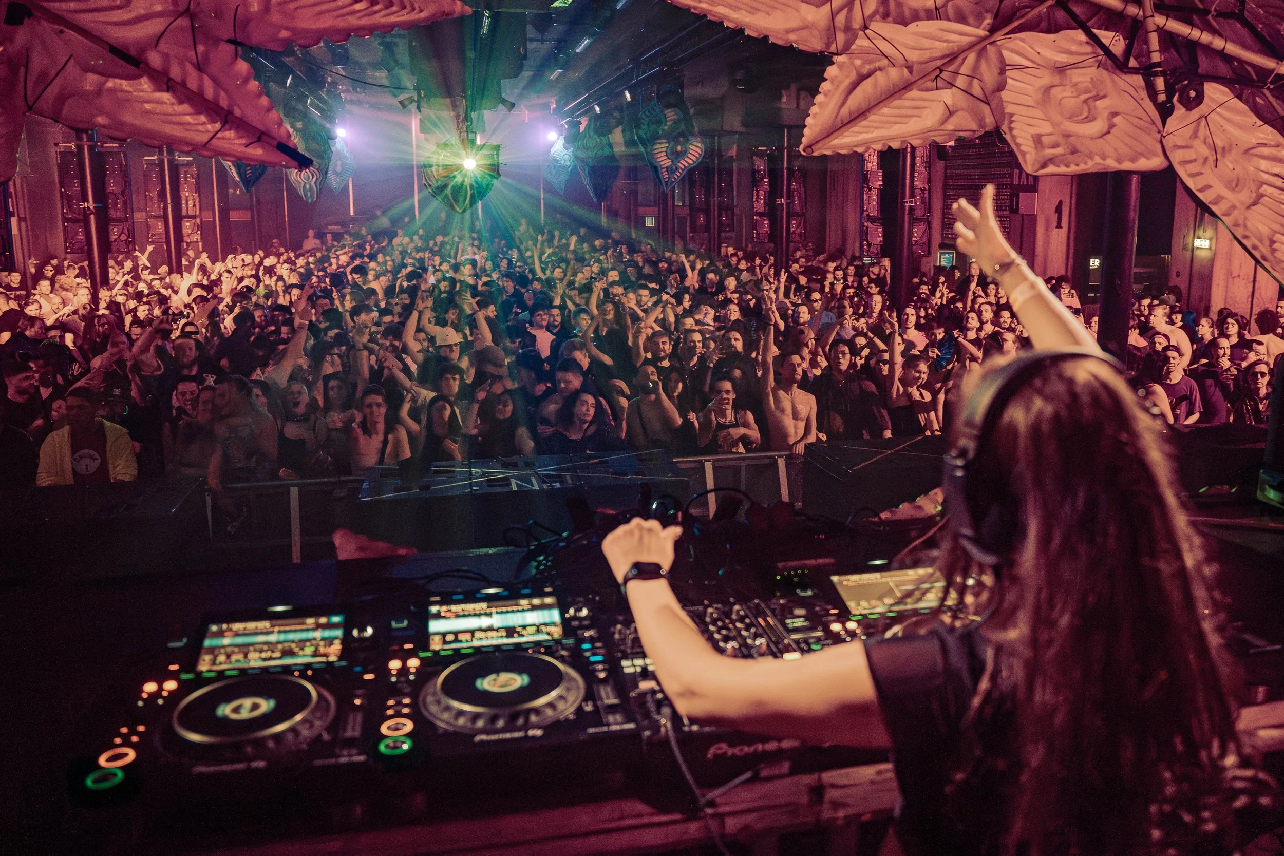 A DJ performing at a nightclub with a large crowd dancing under colorful lighting and decorations.