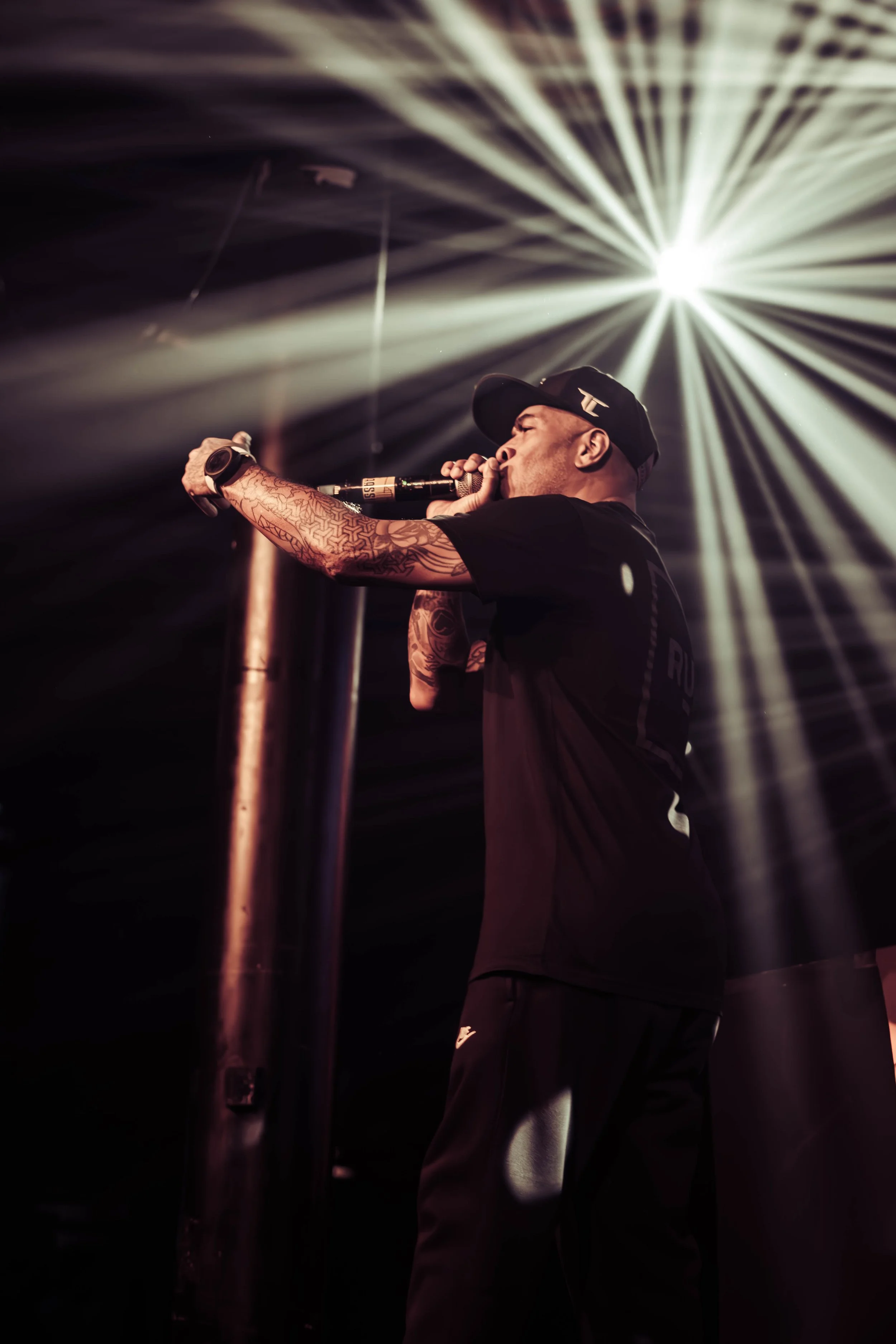 A man wearing a black cap and athletic attire on stage, holding a microphone close to his mouth, with bright stage lights creating a starburst pattern behind him.