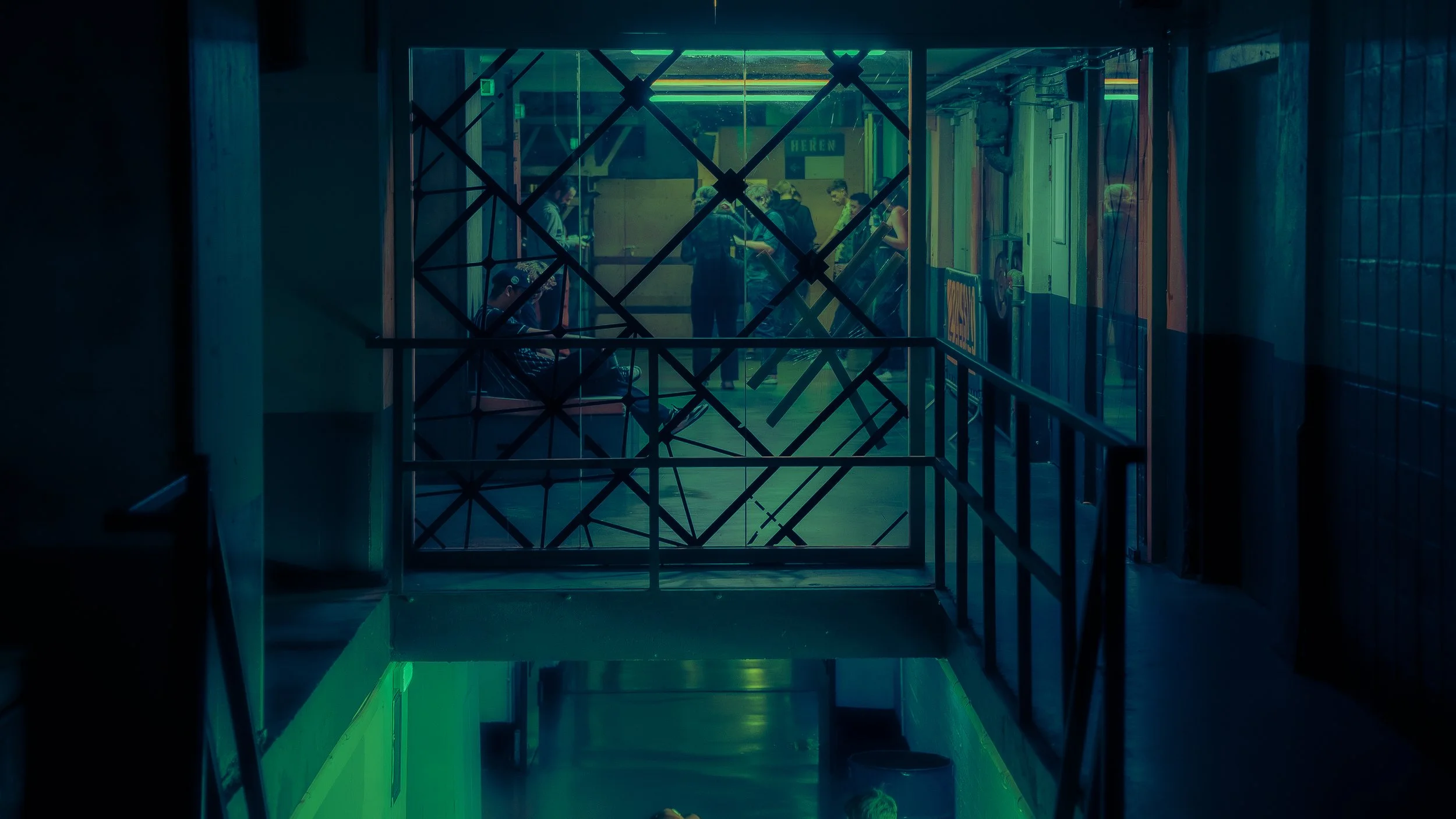 A view through a security gate into a hallway with people waiting and walking, illuminated by colorful neon lights.
