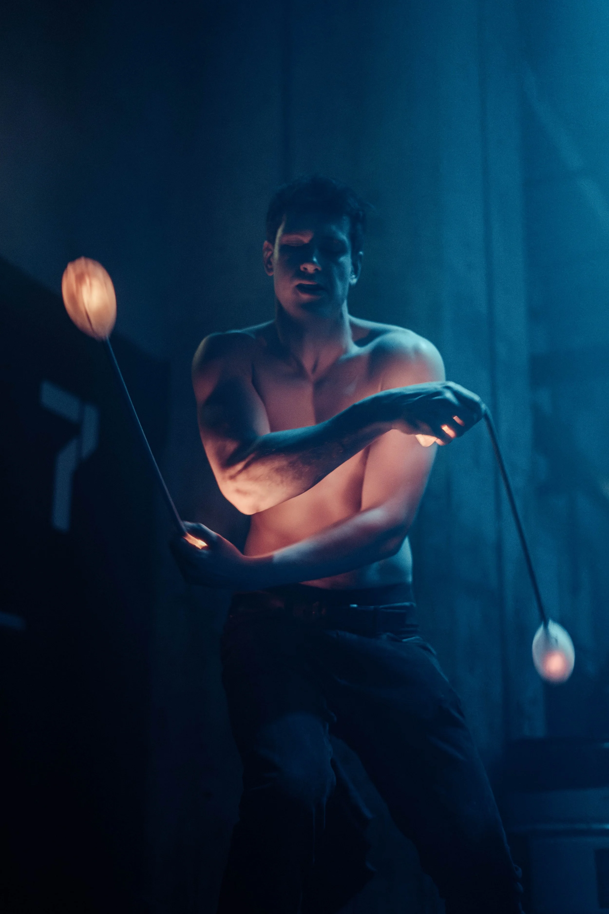 A shirtless young man with dark hair and tattoos on his left arm, holding a microphone and possibly performing on stage with blue lighting.