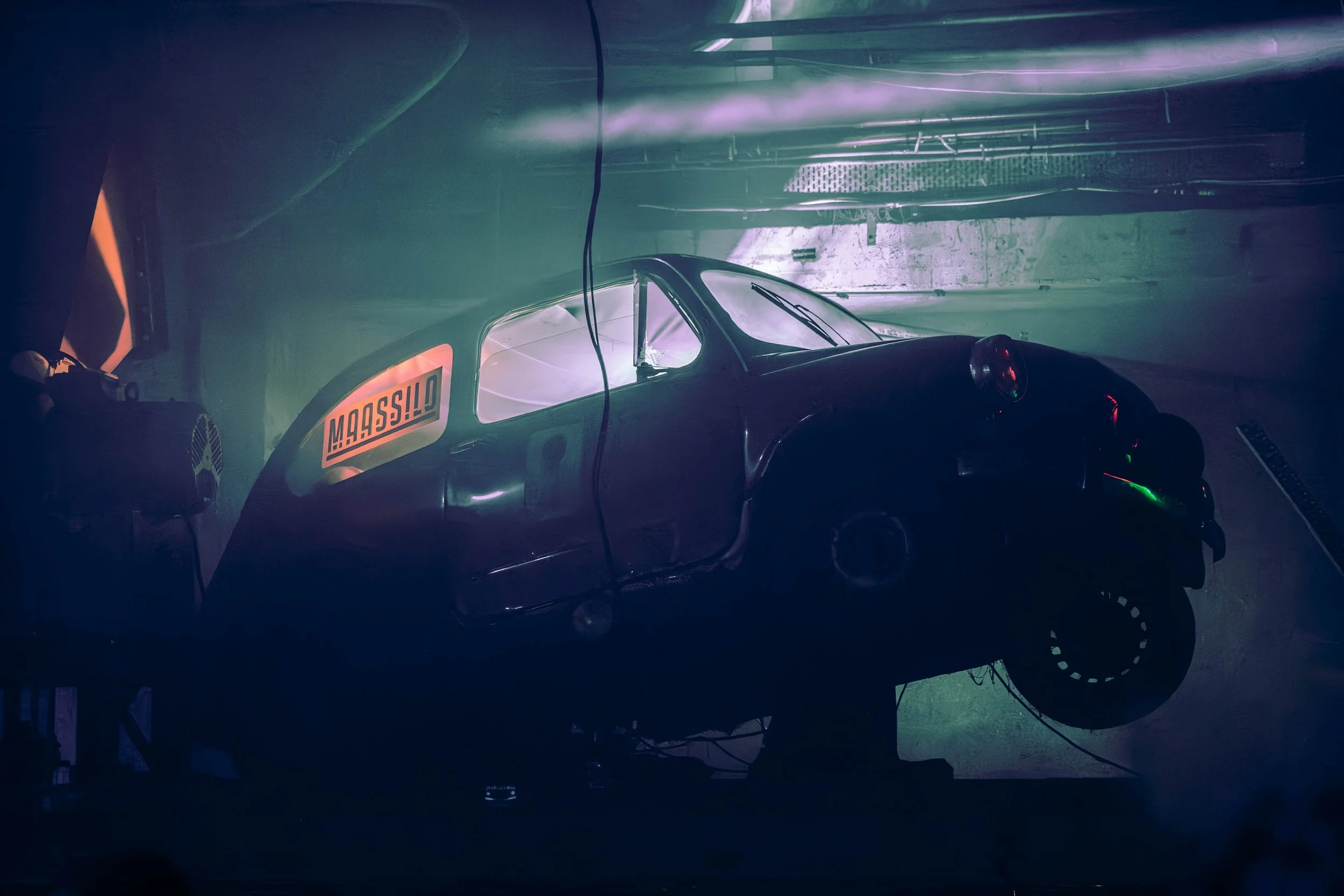 A race car inside a garage with green lighting, a large orange sticker on the side, and various equipment in the background.