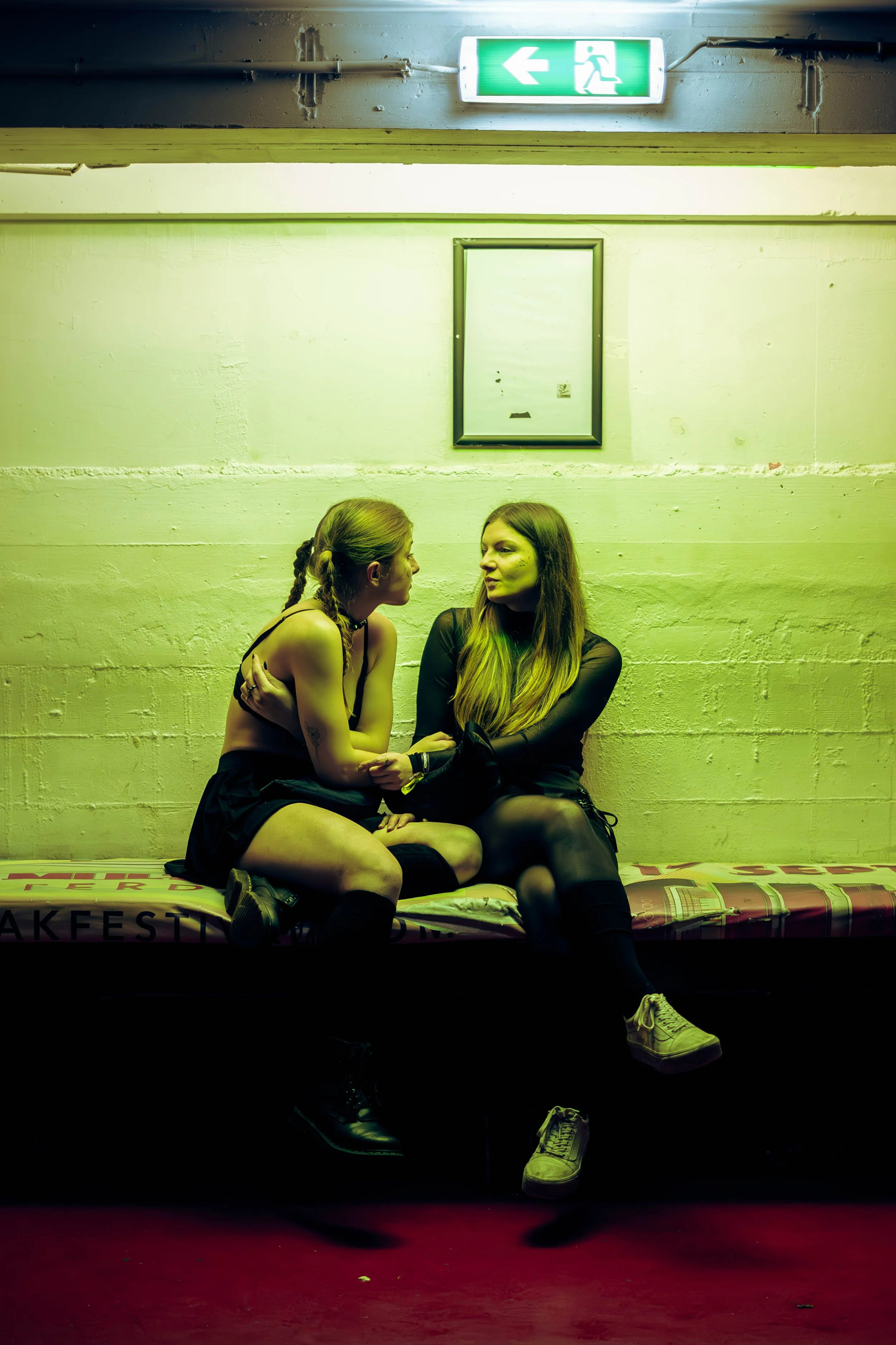 Two women sitting on a bench under a green emergency exit sign, with one woman facing the other and engaging in conversation. The environment appears to be an indoor, possibly underground, area with a red floor and a white wall.