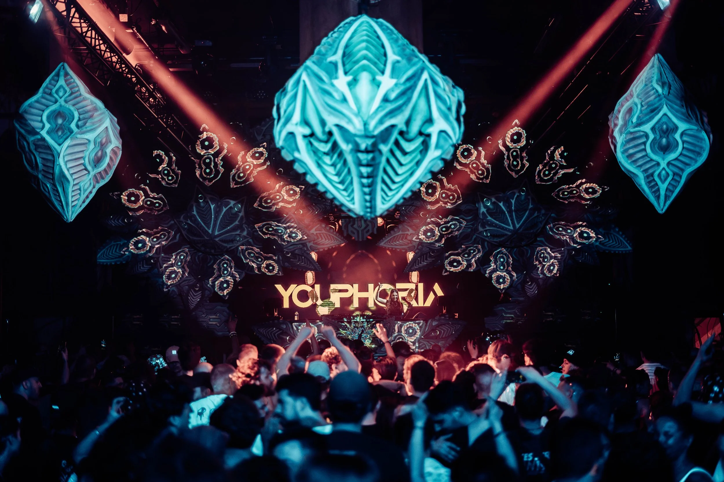 A crowded concert or nightclub with a large illuminated stage featuring geometric patterns and the words 'Y Our PH' displayed. People in the audience are dancing and taking photos, with colorful lights and lasers overhead.