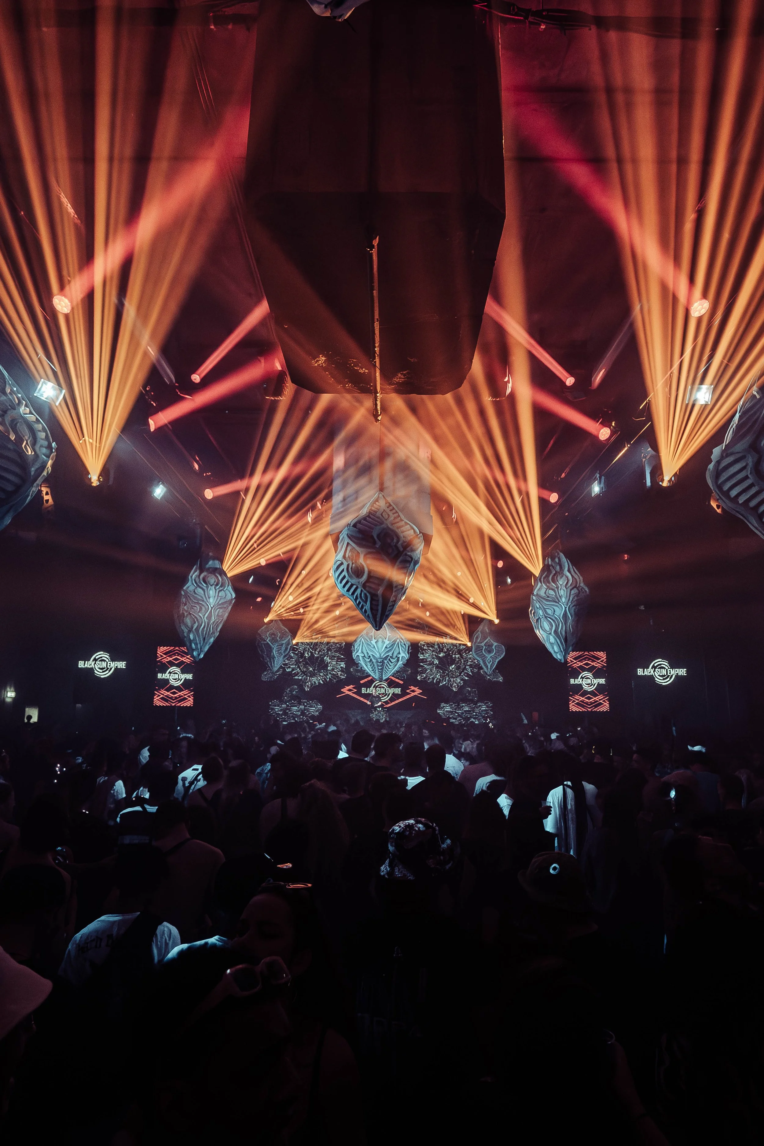 A crowded nightclub or concert with a stage lit by colorful beams of yellow and pink lights, and a DJ booth at the front with the text 'Black Sun Empire' displayed on screens.