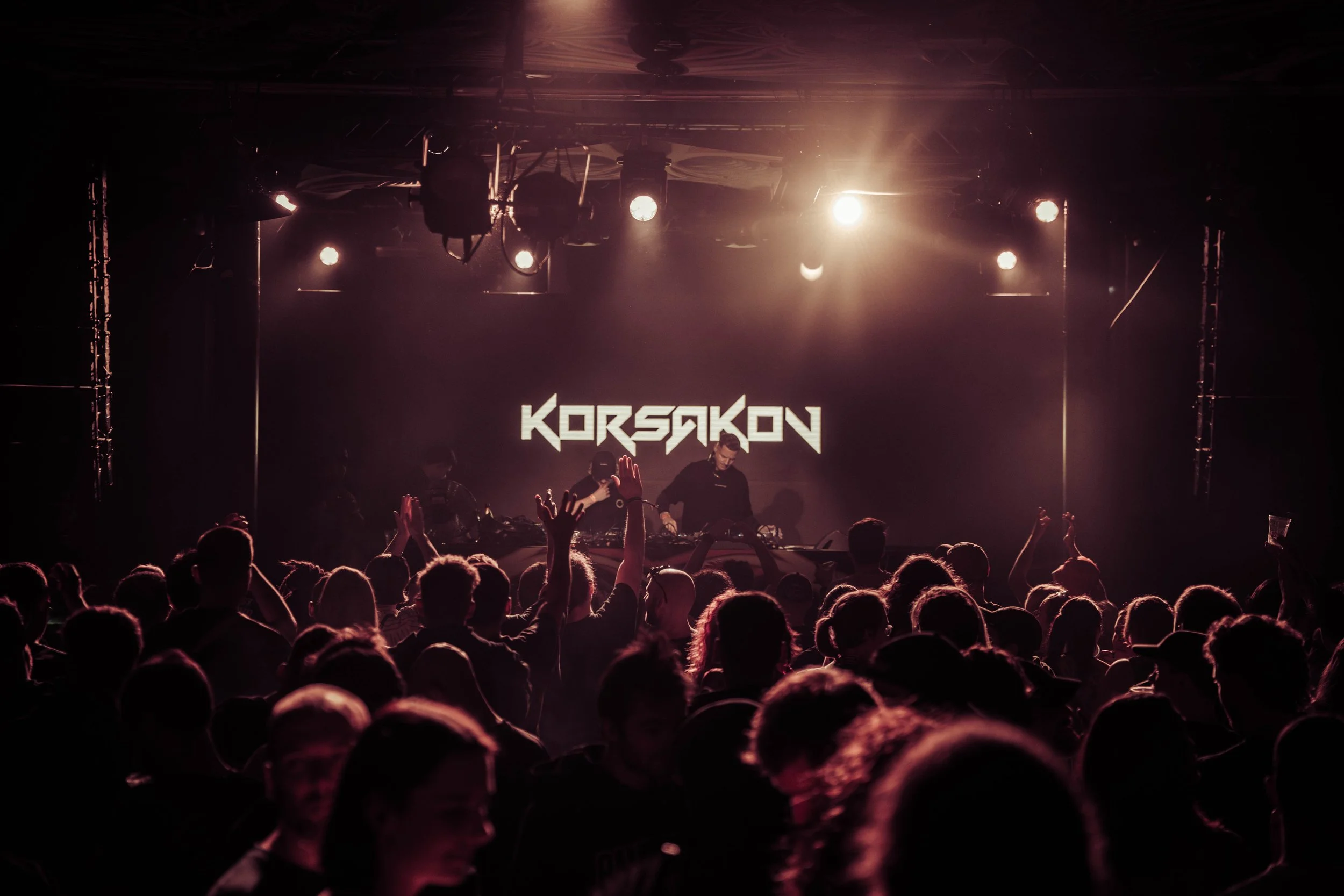 Crowd attending a music concert with stage lights illuminating a DJ performing and a screen displaying the name 'KORSARON'.