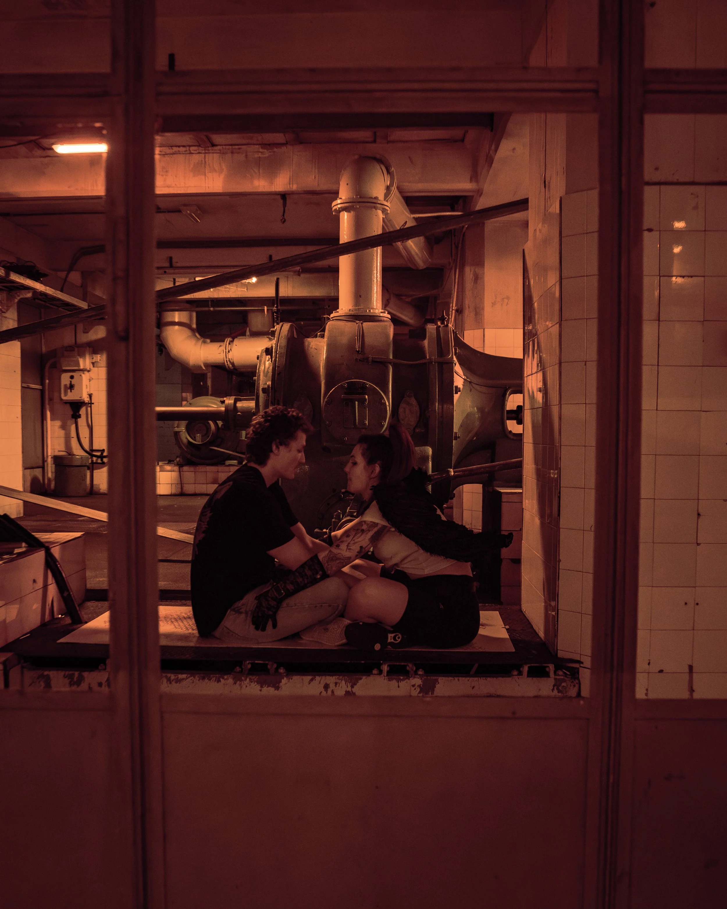 Two people sitting on the floor in front of industrial machinery, engaged in a conversation or activity, framed by metal bars and a tiled wall.