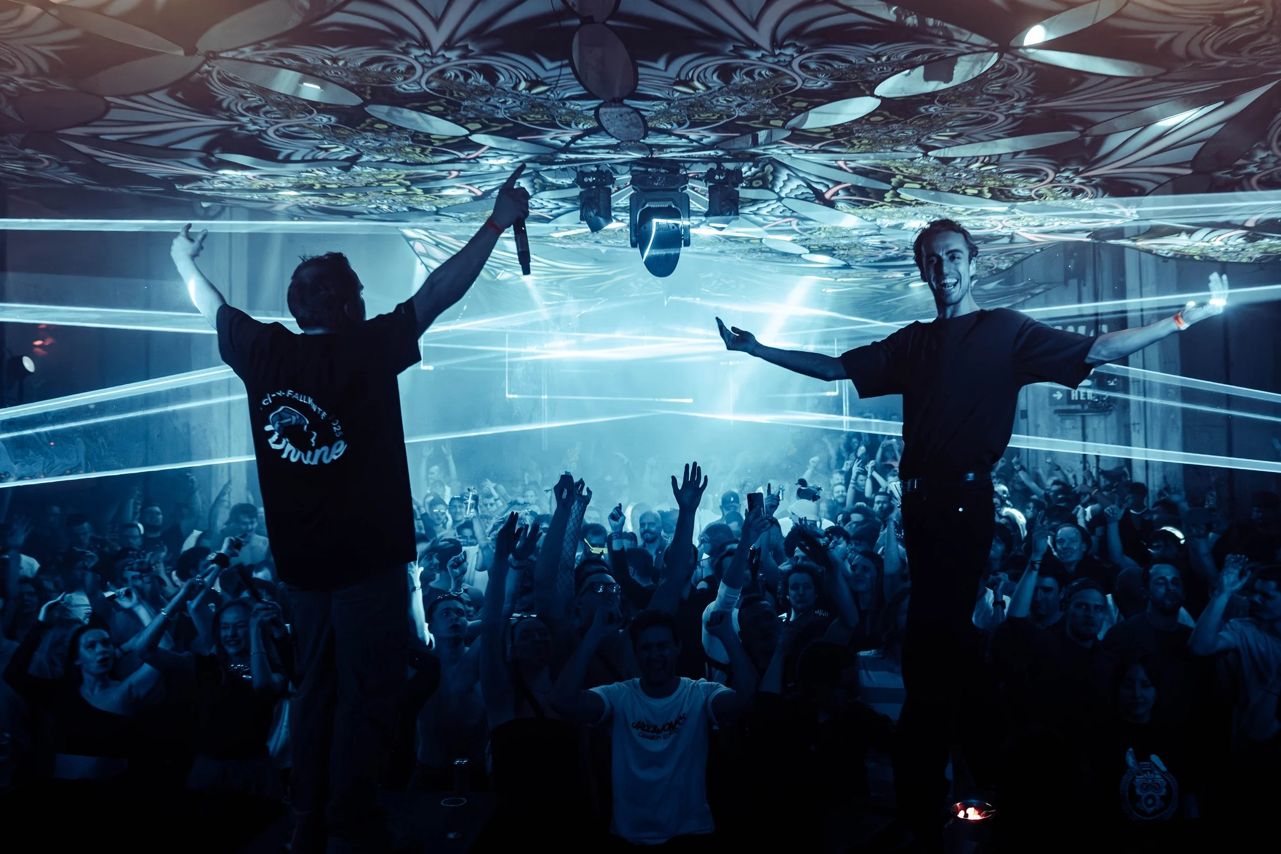 A lively music concert with a large crowd dancing and enjoying the performance. Two performers are center stage; one is seen from the back with arms raised, and the other is smiling with arms outstretched. The stage features decorative lighting and a