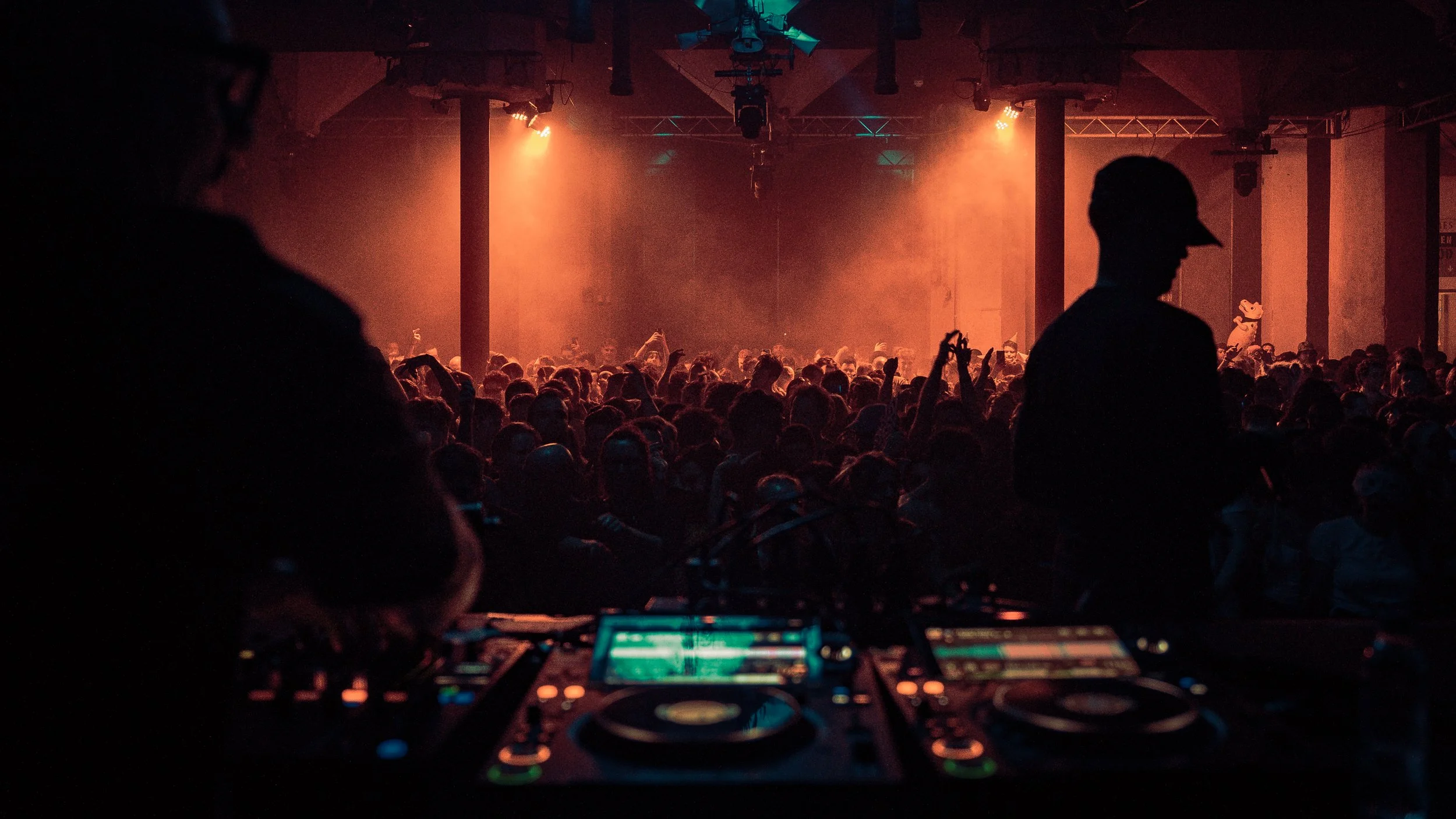 A DJ setup with a mixing console in the foreground, silhouettes of people dancing and enjoying a concert or music event in the background with warm orange lighting.