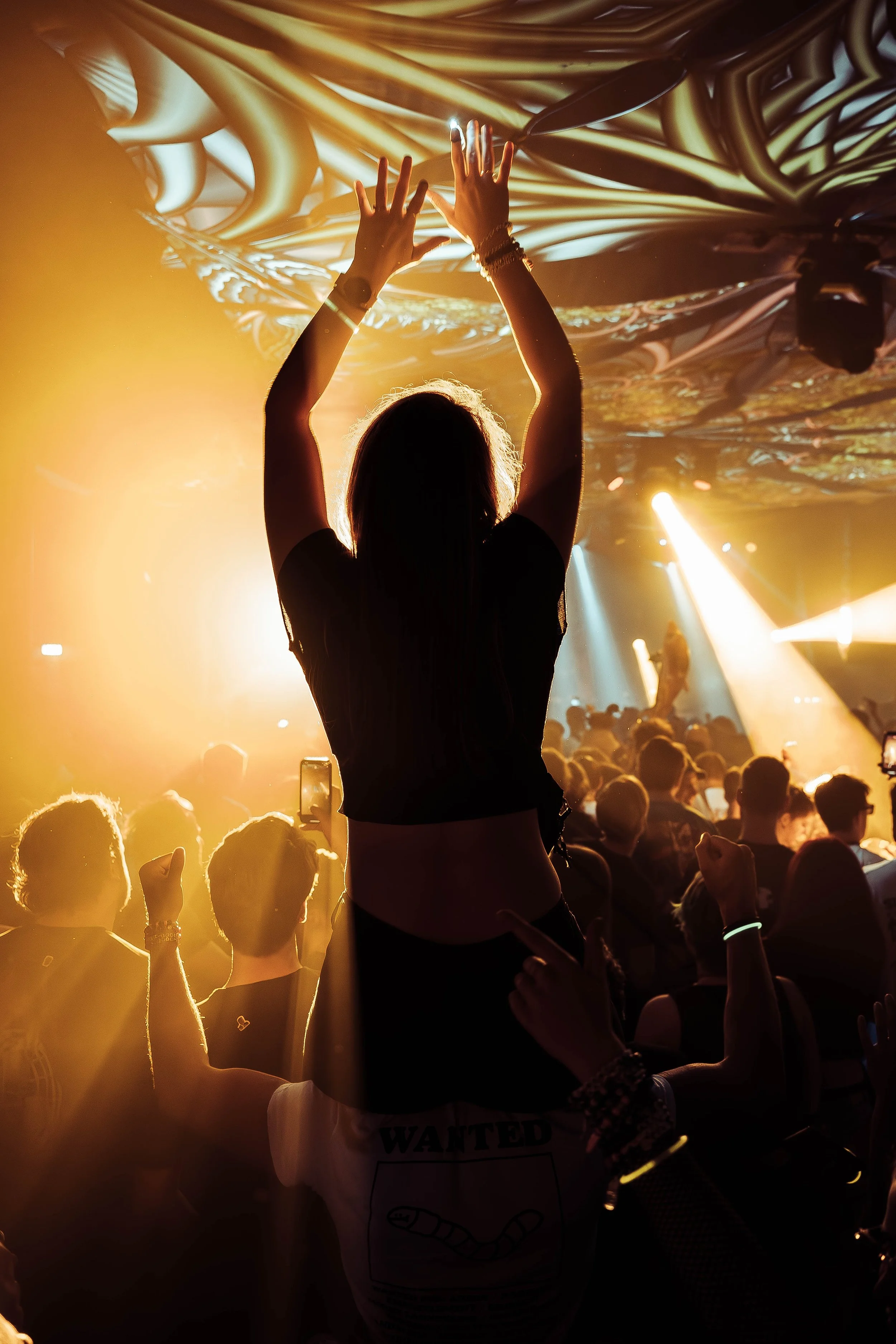 A person with raised hands at a concert or nightclub with colorful, swirling lights on the ceiling and an audience in the background.