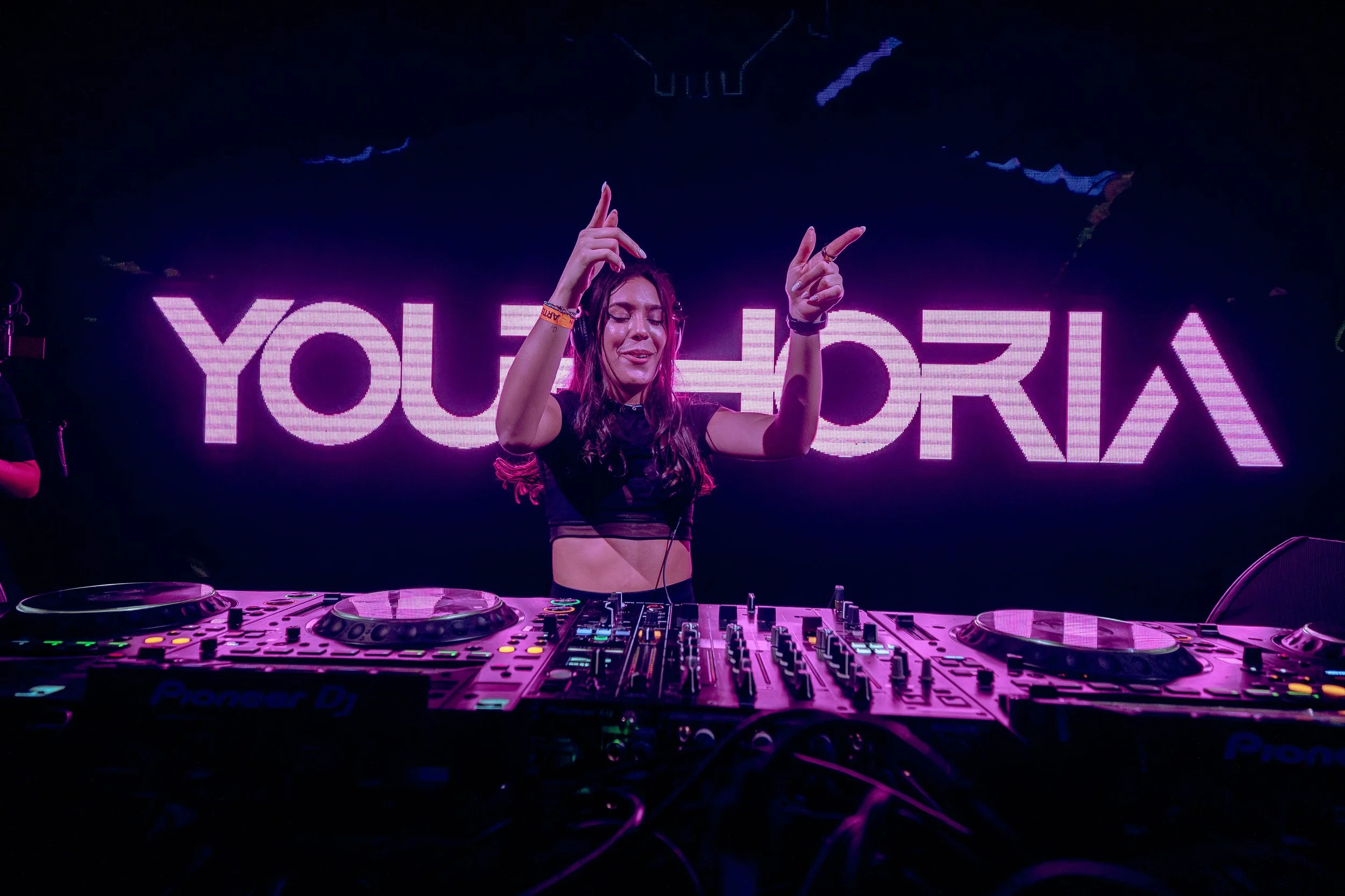 Female DJ performing at a nightclub with neon lights in the background that spell out a word, wearing headphones and making hand gestures, with DJ equipment in front of her.