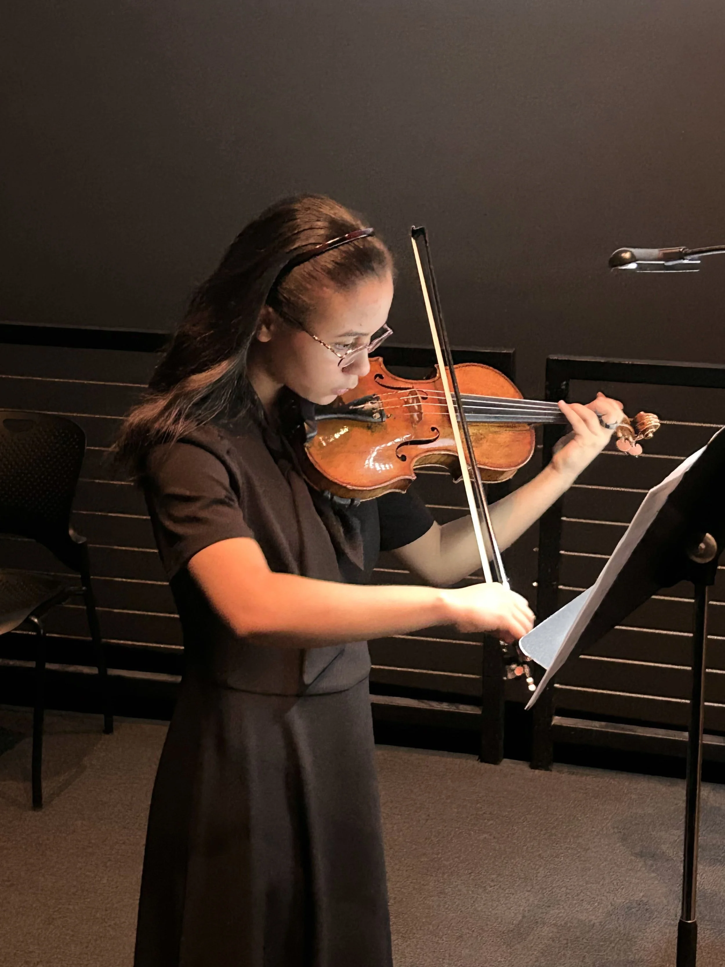 A woman wearing glasses playing the violin while reading sheet music in a dimly lit room.
