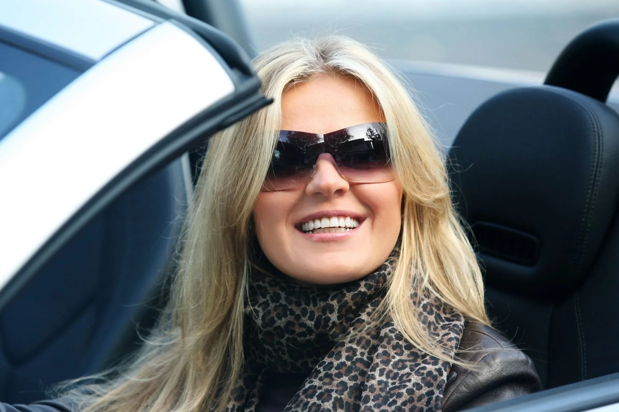 A woman with long blonde hair wearing large sunglasses and a leopard print scarf smiling in the backseat of a car.