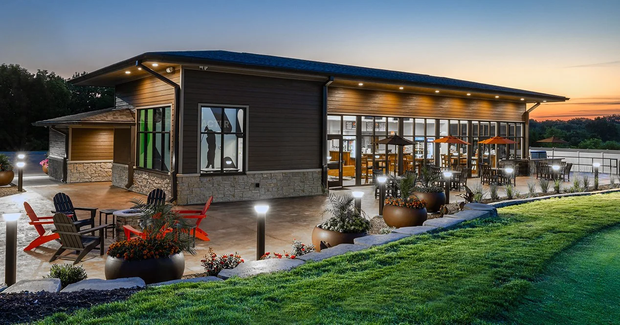 Exterior of a modern golf club house with large windows and patio seating, landscaped with potted plants and grass, during sunset with outdoor lighting.
