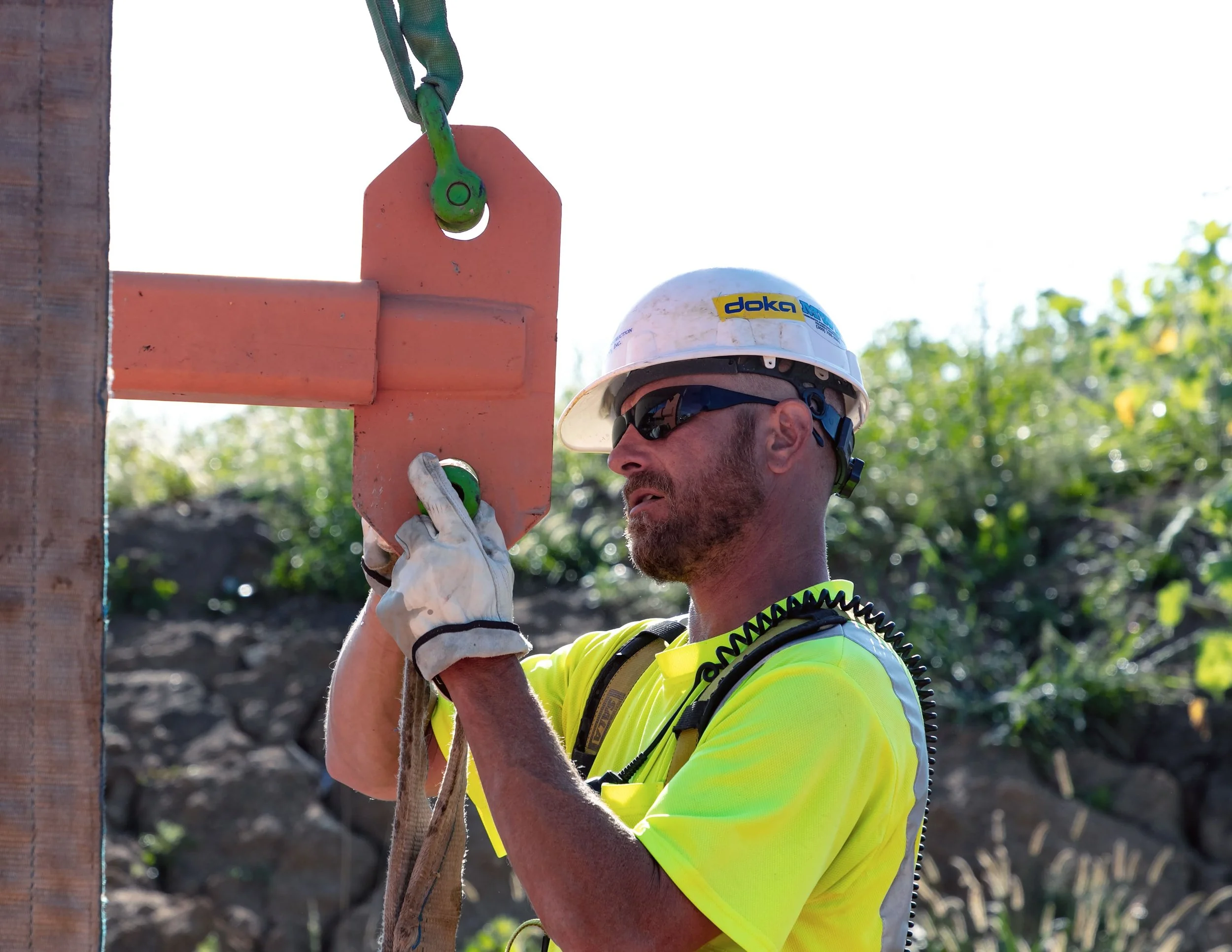 A construction worker wearing a white safety helmet with a yellow and blue sticker, sunglasses, a high-visibility yellow shirt, and work gloves, is at a construction site adjusting or attaching a wooden beam with a large green metal hook.