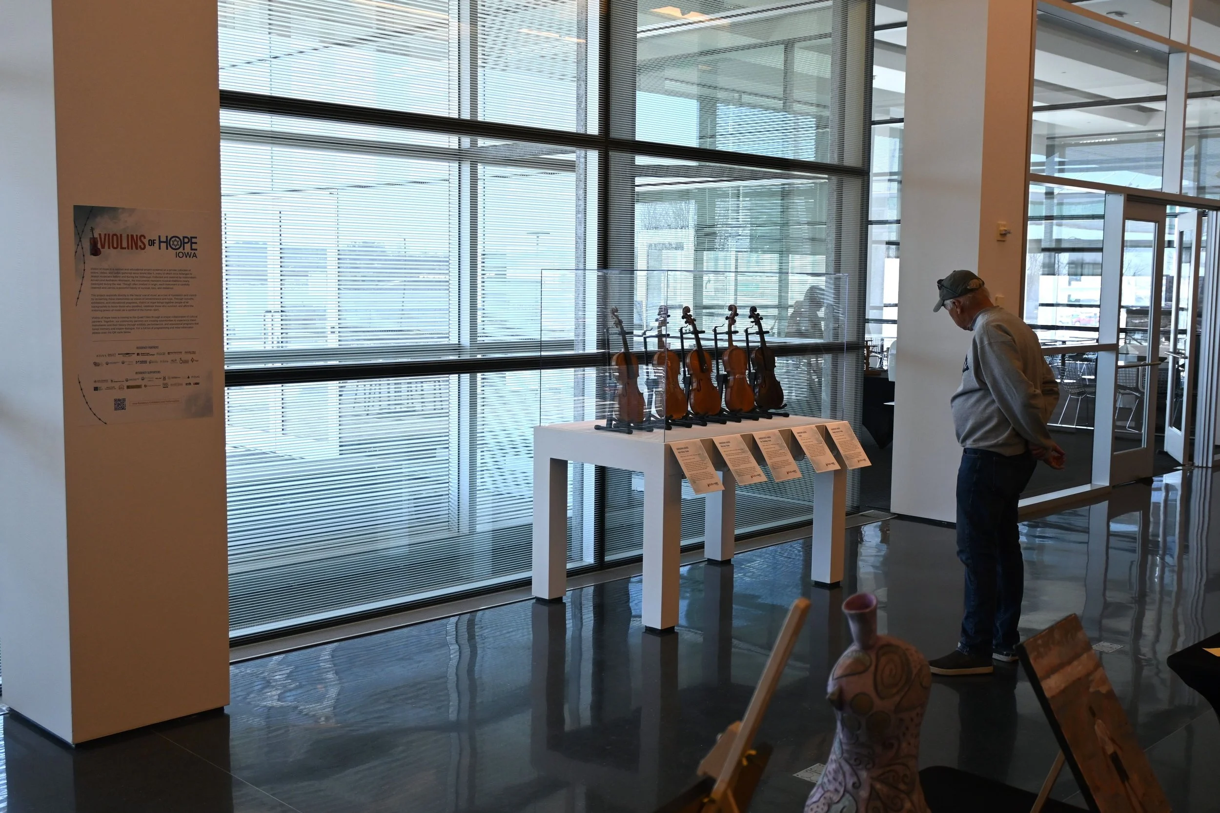 Inside a modern building with large windows and glass walls, a man wearing a gray hoodie and cap looks at display tables with informational papers, with violins displayed on a stand nearby, and a poster on the wall about Violins of Hope Iowa.