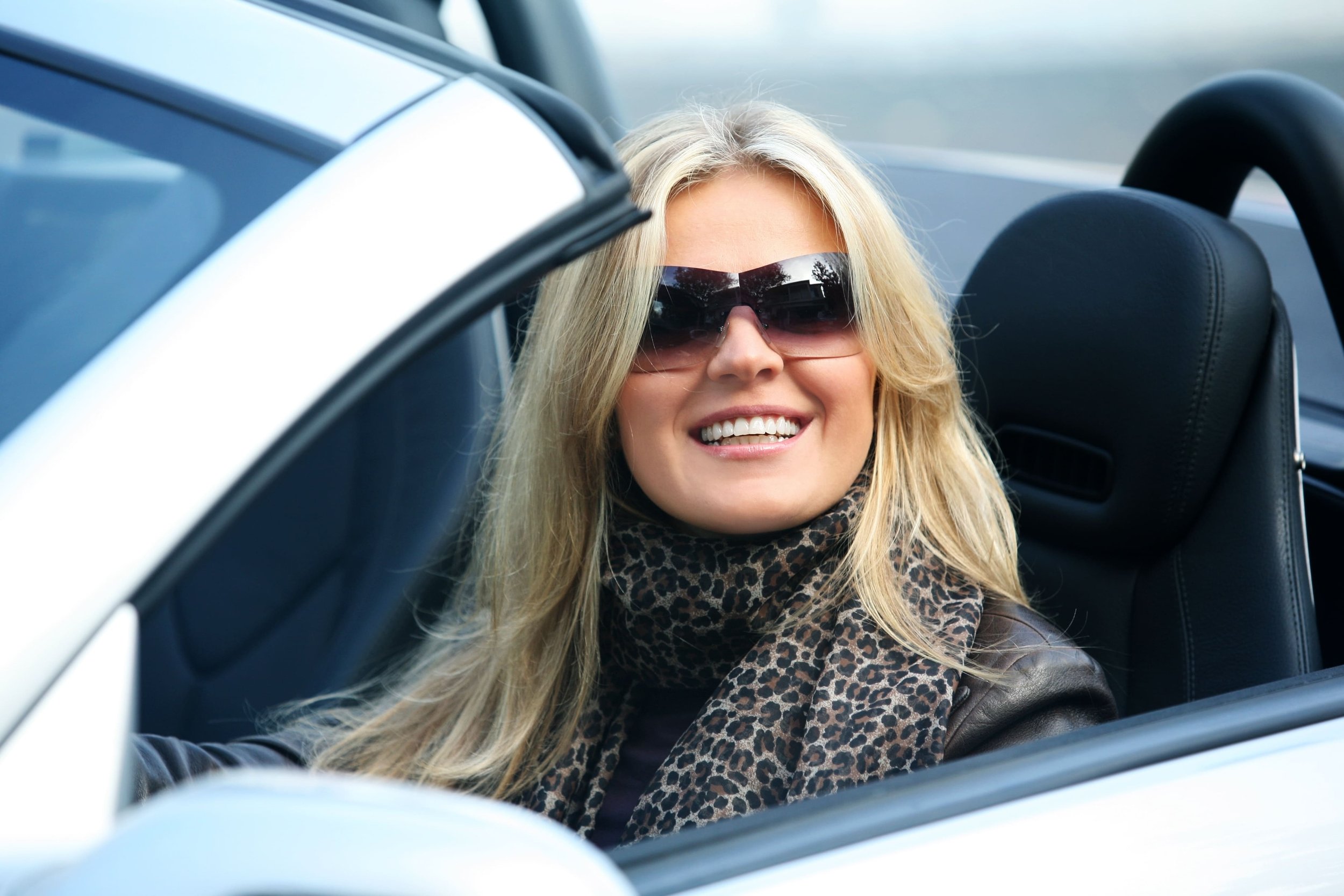 A woman with blonde hair smiling and wearing sunglasses while sitting in a convertible car.