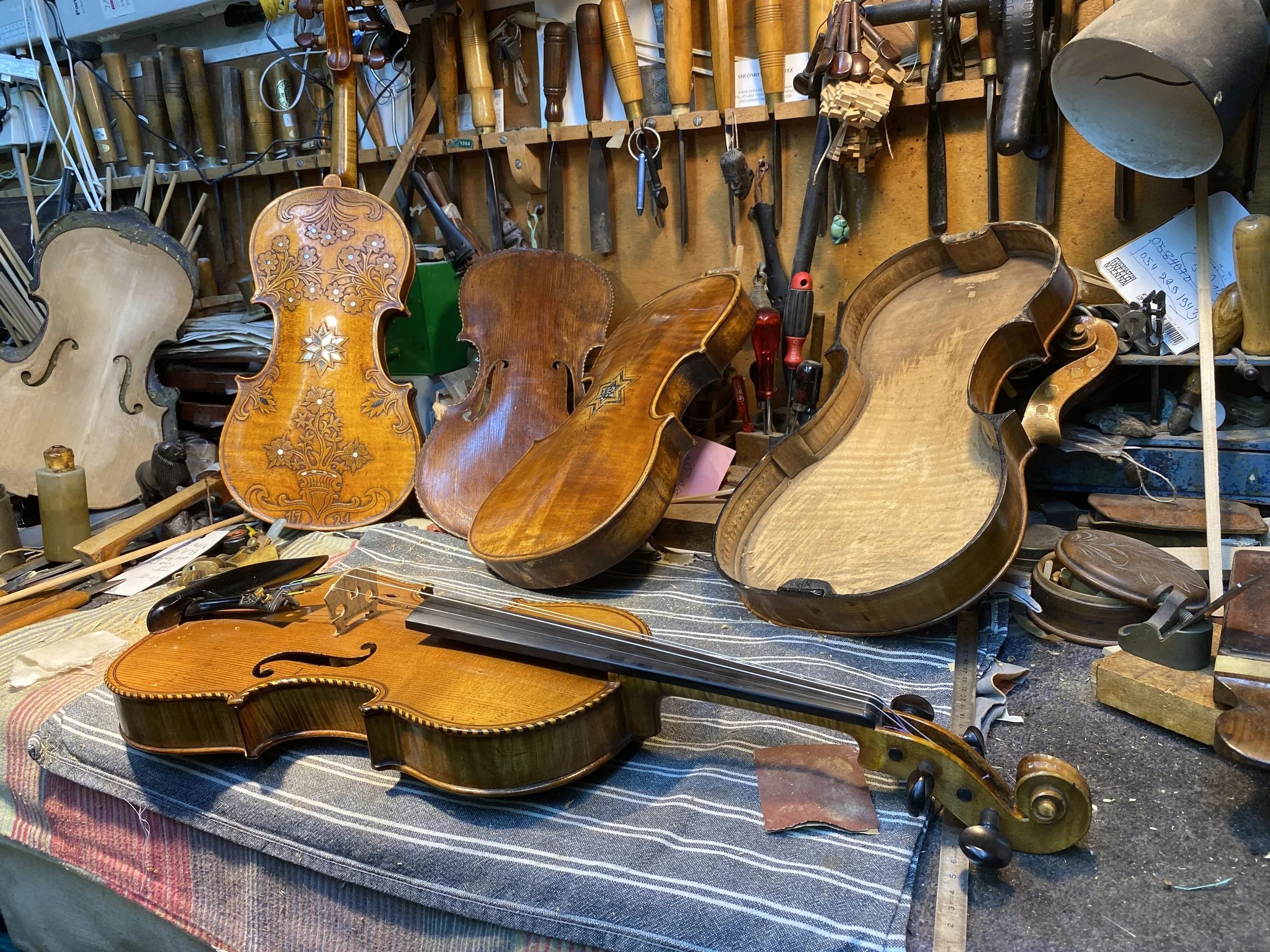A cluttered workbench with various woodworking tools hanging on a wooden wall, and several violins in different stages of repair or construction placed on the table and leaning against the wall.