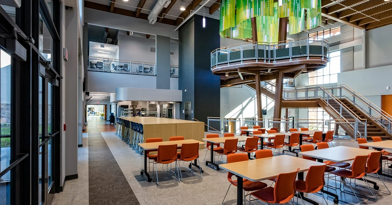 Modern indoor space with orange chairs arranged around tables, a large central tree-like structure with colorful green hanging art, and a staircase leading to an upper level with glass-walled offices.