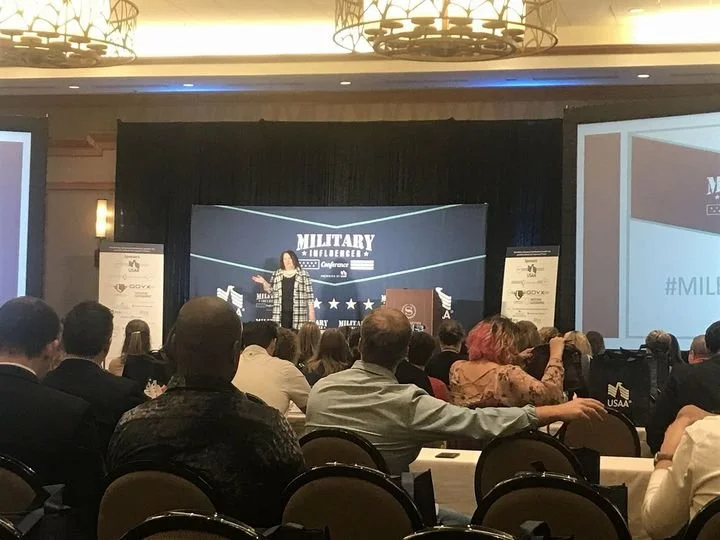 A woman speaking on stage at the Military Intelligence Conference with audience members seated in front, and large screens displaying the conference branding in the background.